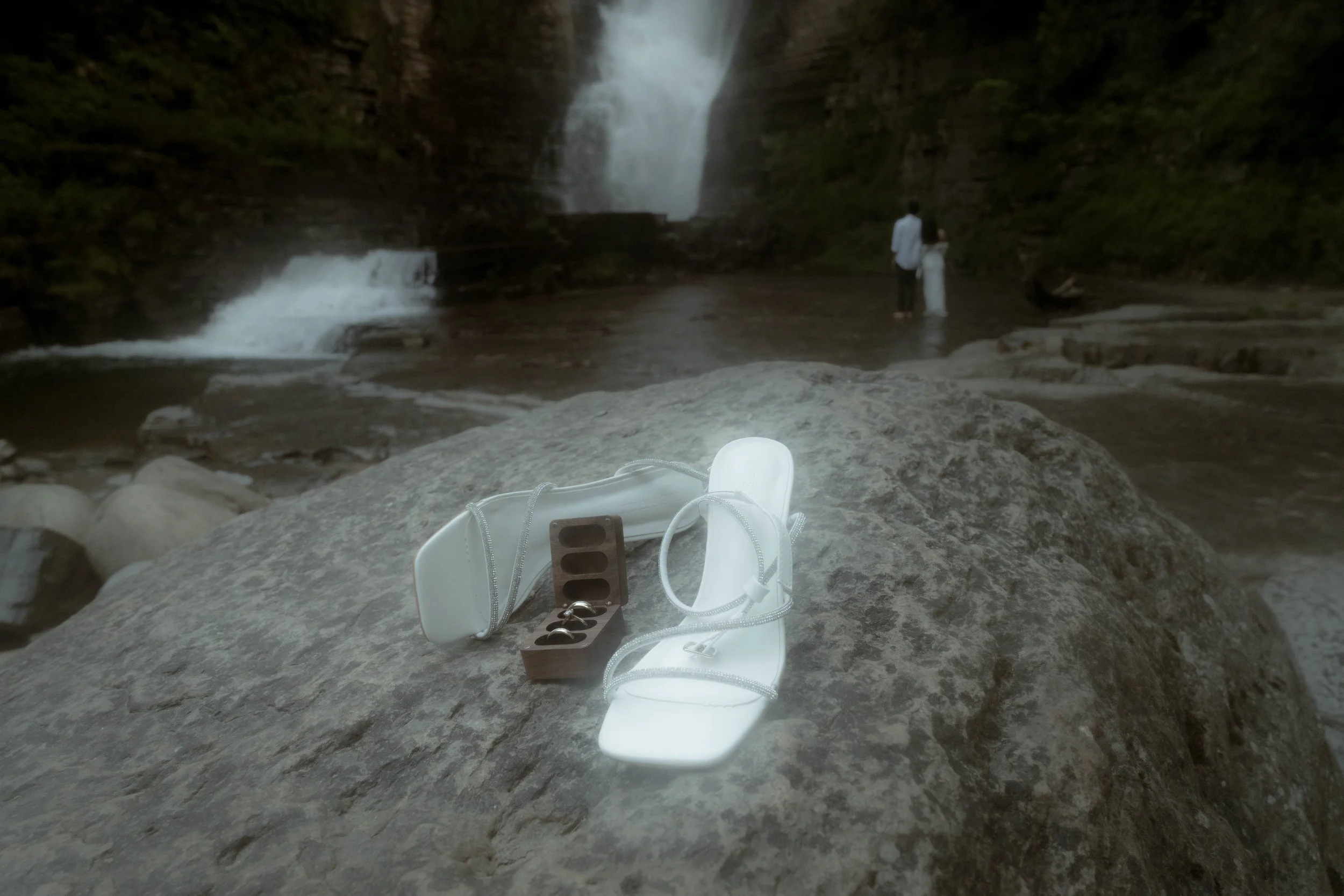 Chaussures de randonnée, lunettes de soleil et appareil photo posés sur une roche en face d'une cascade dans la nature.