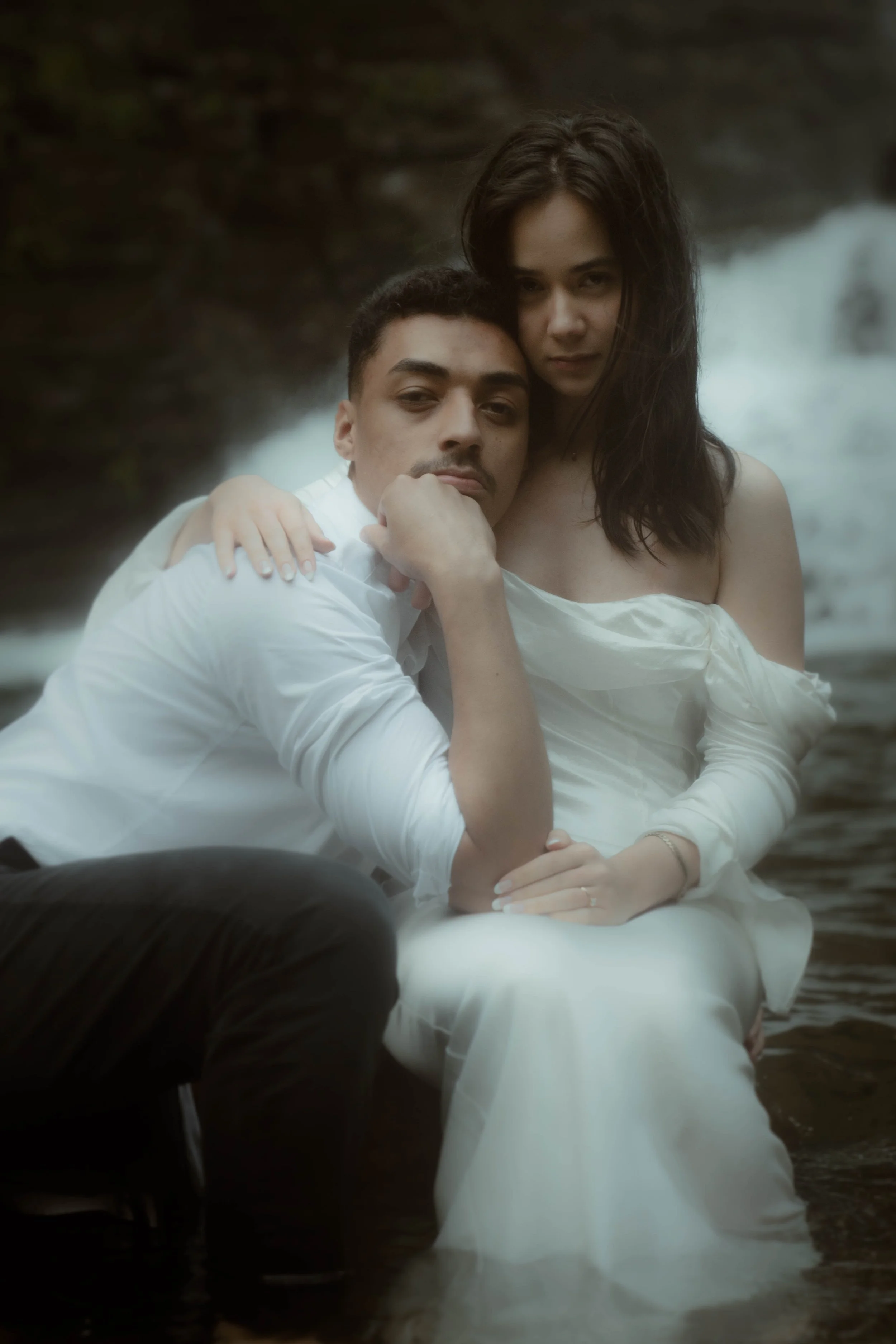 Deux personnes en robe blanche se tiennent près d'une rivière ou d'une cascade, en pleine nature, lors d'une séance photo romantique.