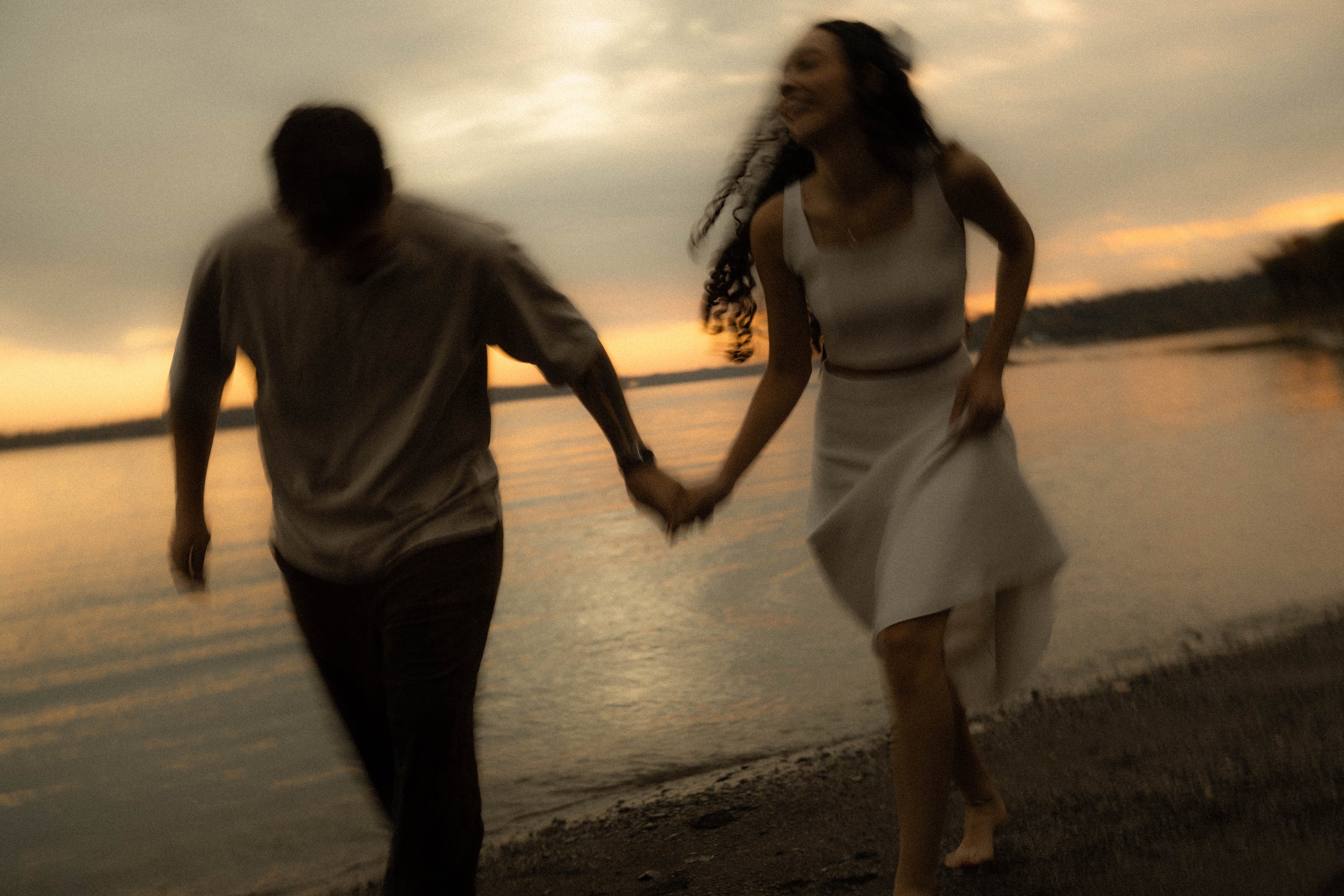 Un couple se promène main dans la main au bord de la mer au coucher du soleil, souriants et heureux.