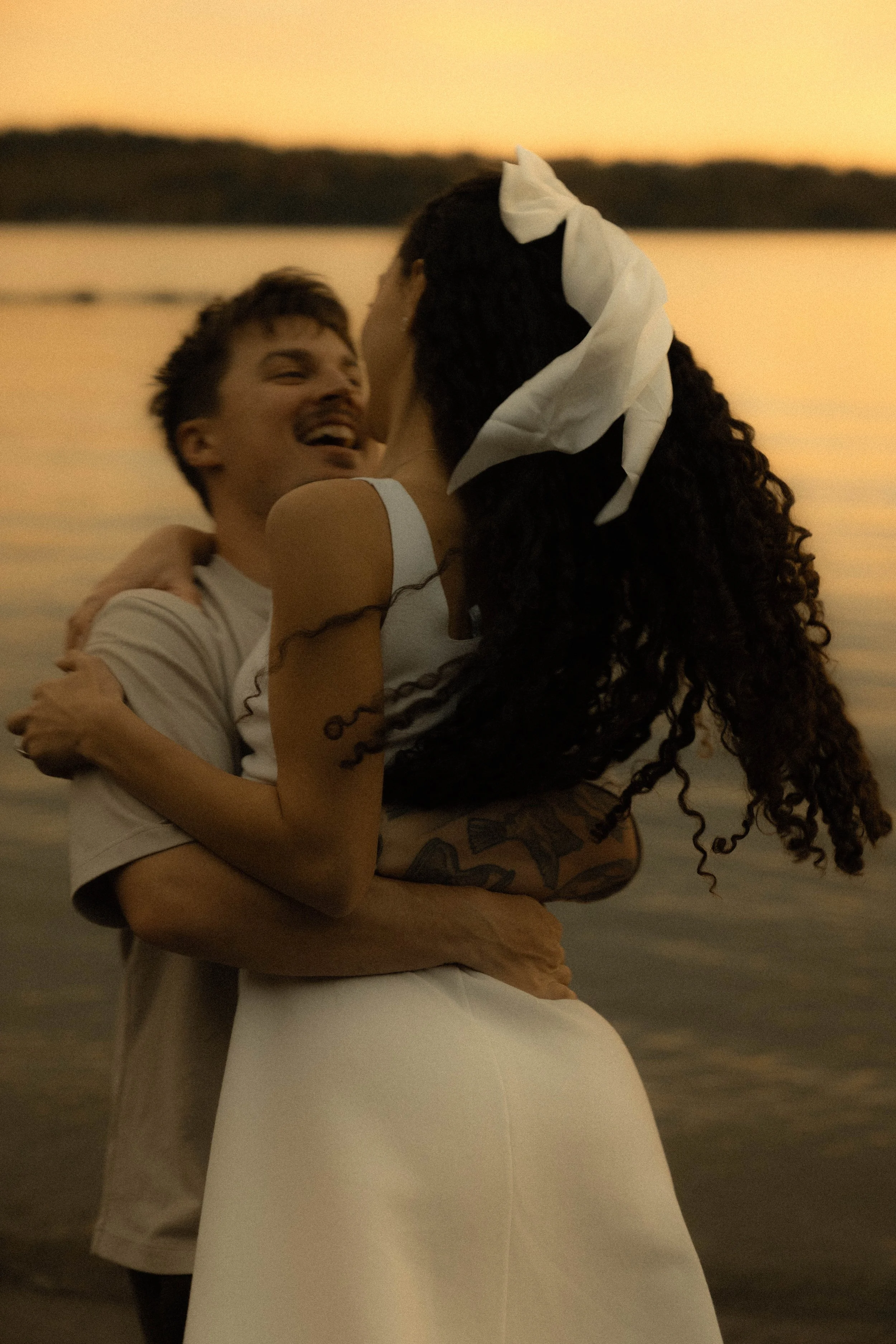 Un couple danse près de la mer au coucher du soleil, souriant et en train de se tenir dans une étreinte.
