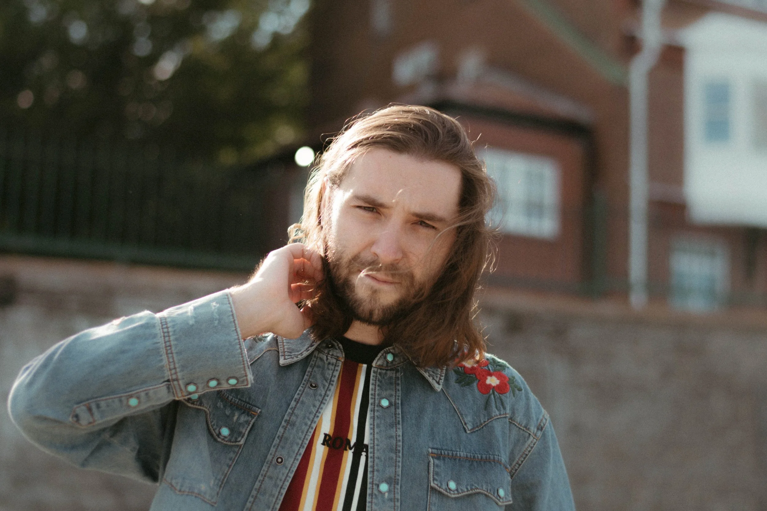 Jeune homme avec long hair et barbe, portant une veste en jean avec des broderies de fleurs rouges et un t-shirt rayé, se tenant dehors en regardant l'appareil photo.