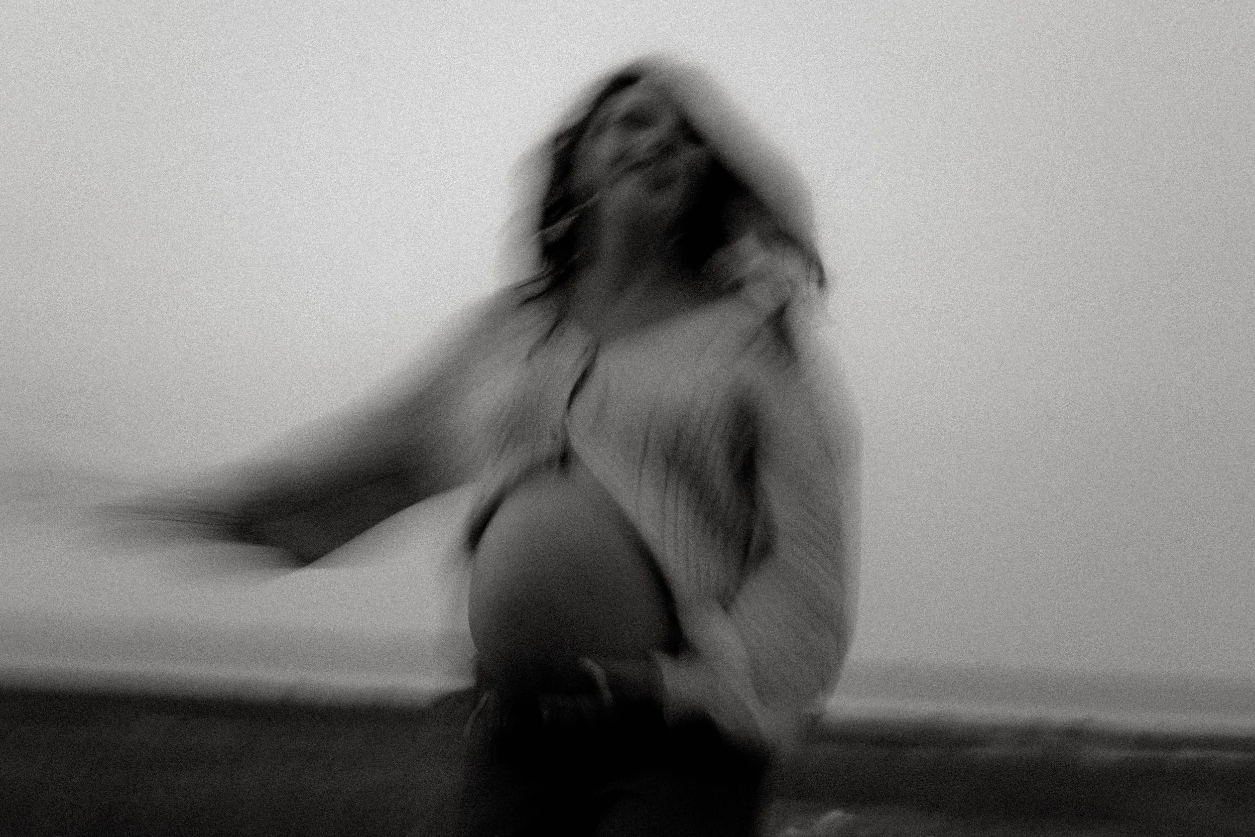 Une personne avec des cheveux longs et bouclés, vue de dos, assise dans une pièce avec un mur blanc, en noir et blanc.