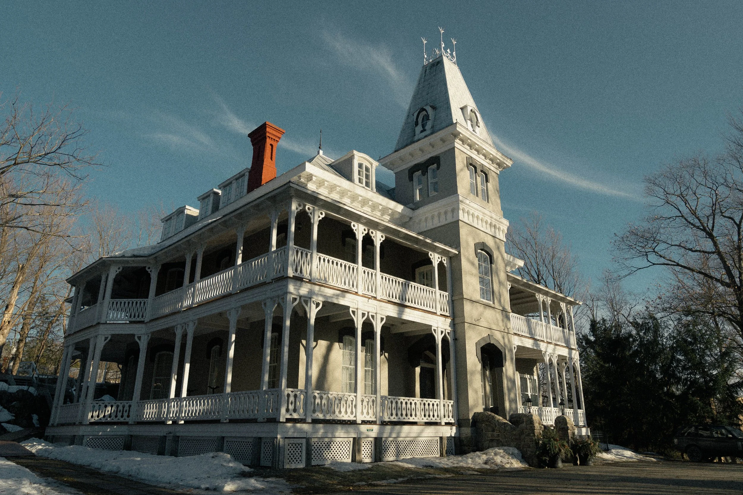 Une grande maison victorienne blanche avec plusieurs étages et une galerie entourée d'une barrière en bois. La maison possède une tour avec un toit pointu et des fenêtres à volets noirs. Il y a des arbres sans feuilles autour de la maison, et il semble faire froid avec des traces de neige sur le sol.