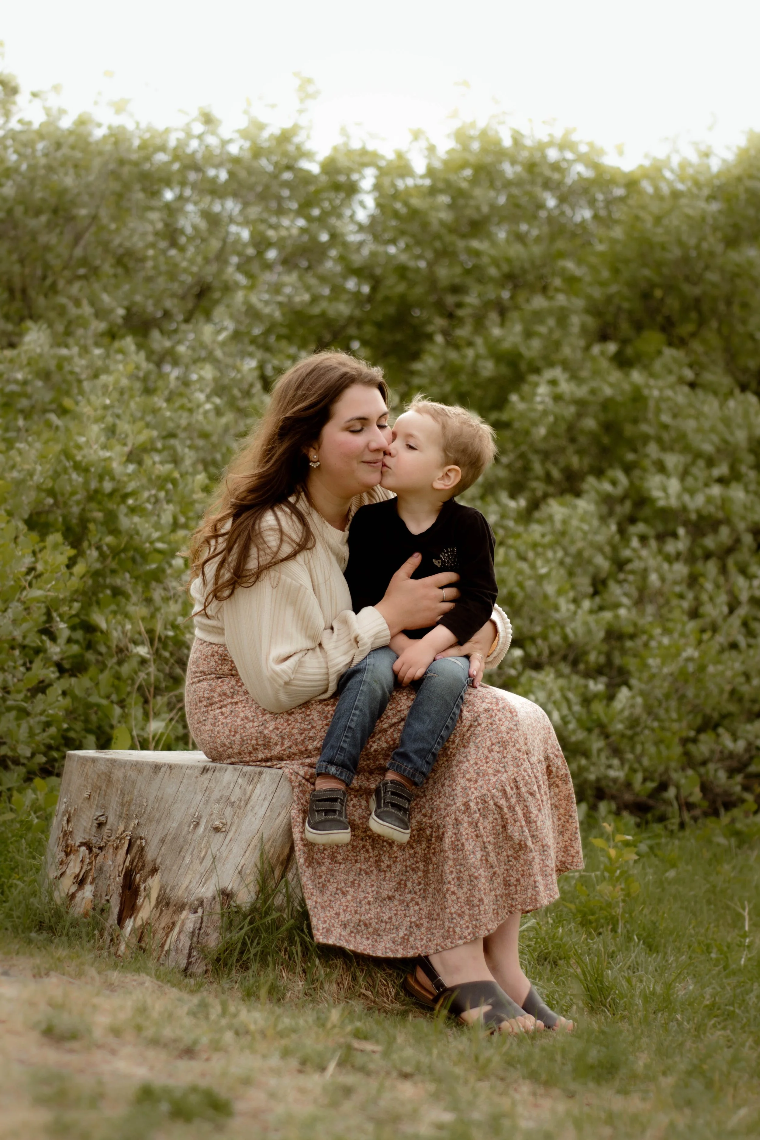 Une femme et un jeune garçon assis sur un tronc d'arbre dans la nature, partageant un câlin et un baiser affectueux, avec des arbres en arrière-plan.