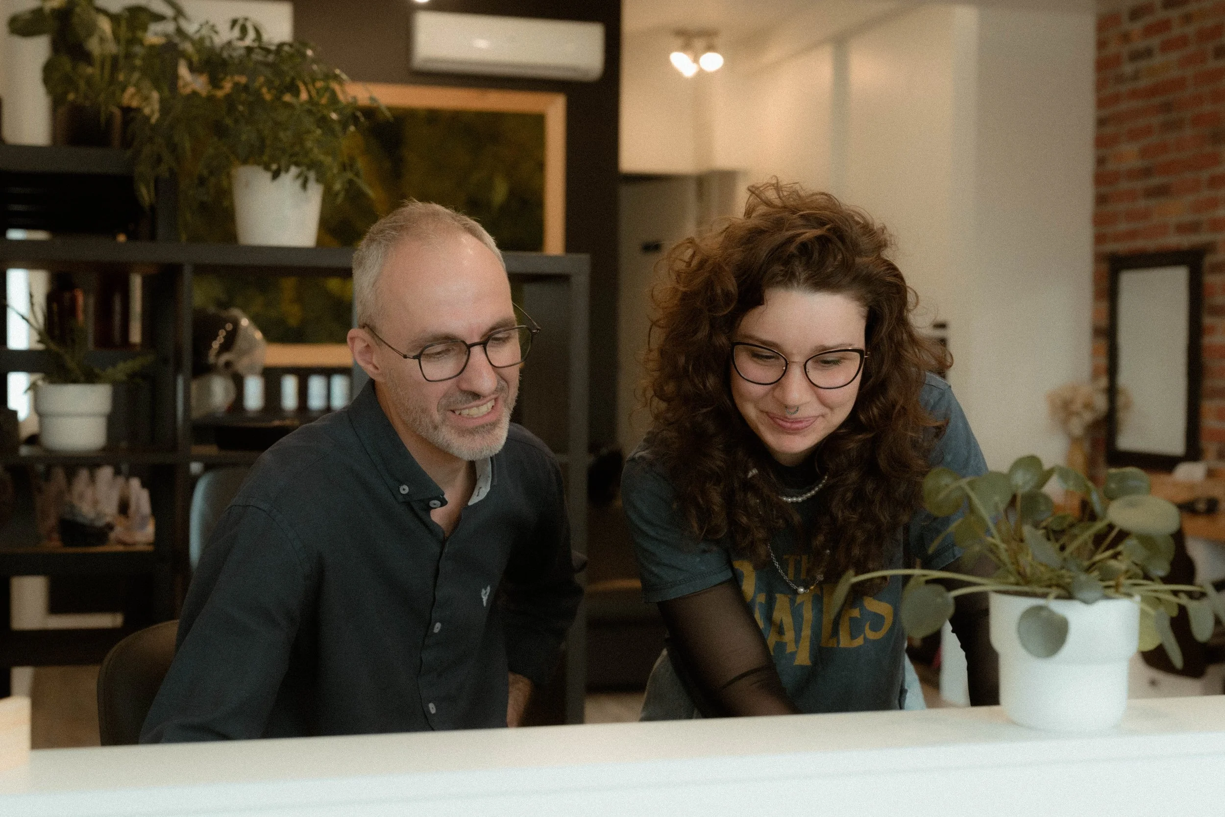 Deux personnes, un homme et une femme portant des lunettes, sont assises à une table et regardent quelque chose, avec des plantes en pot devant eux dans un intérieur chaleureux.