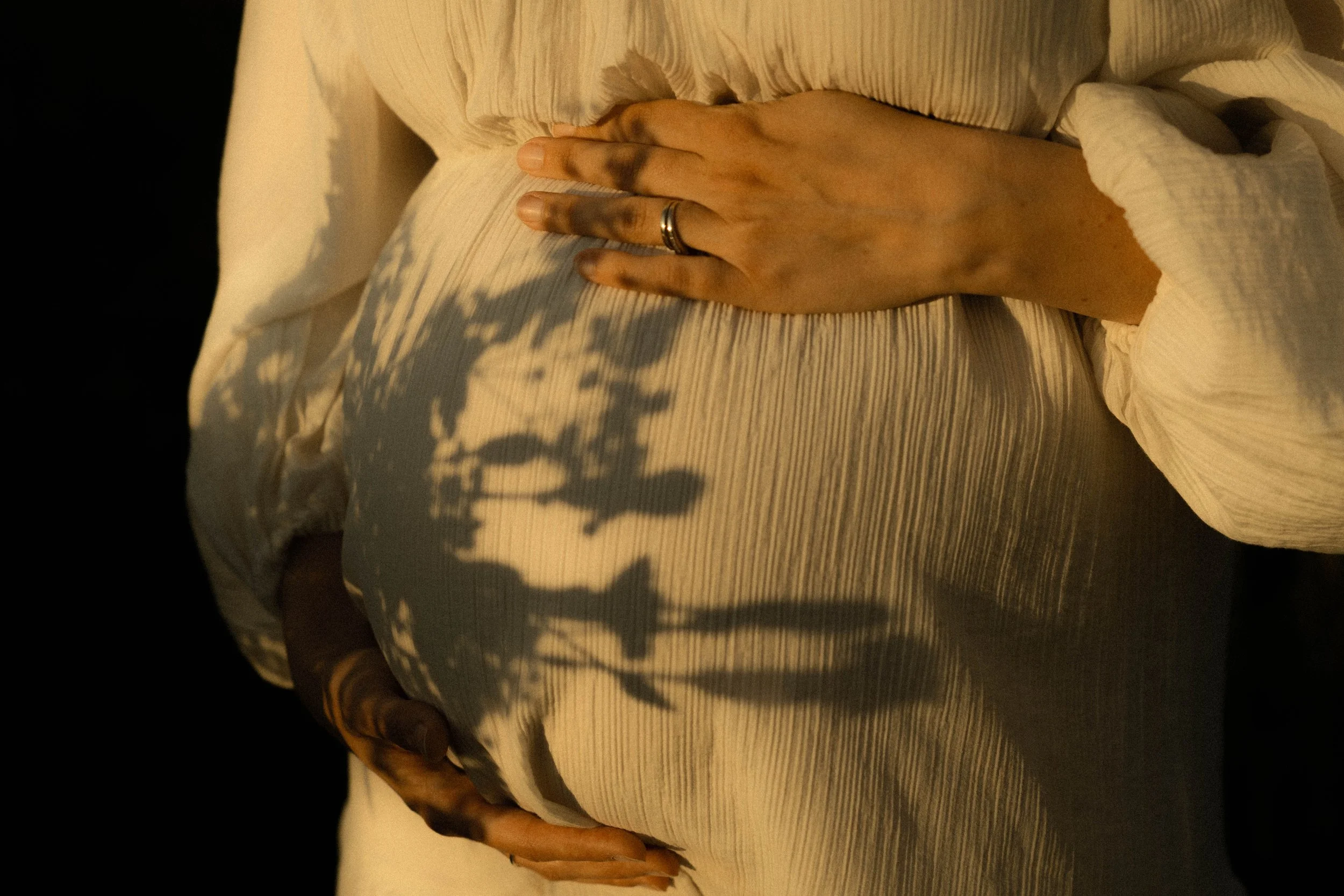 Une main posée sur un lit avec une couverture blanche rayée, shadows de branches d'arbres projetés sur le lit et la main, une bague à l'annulaire. 