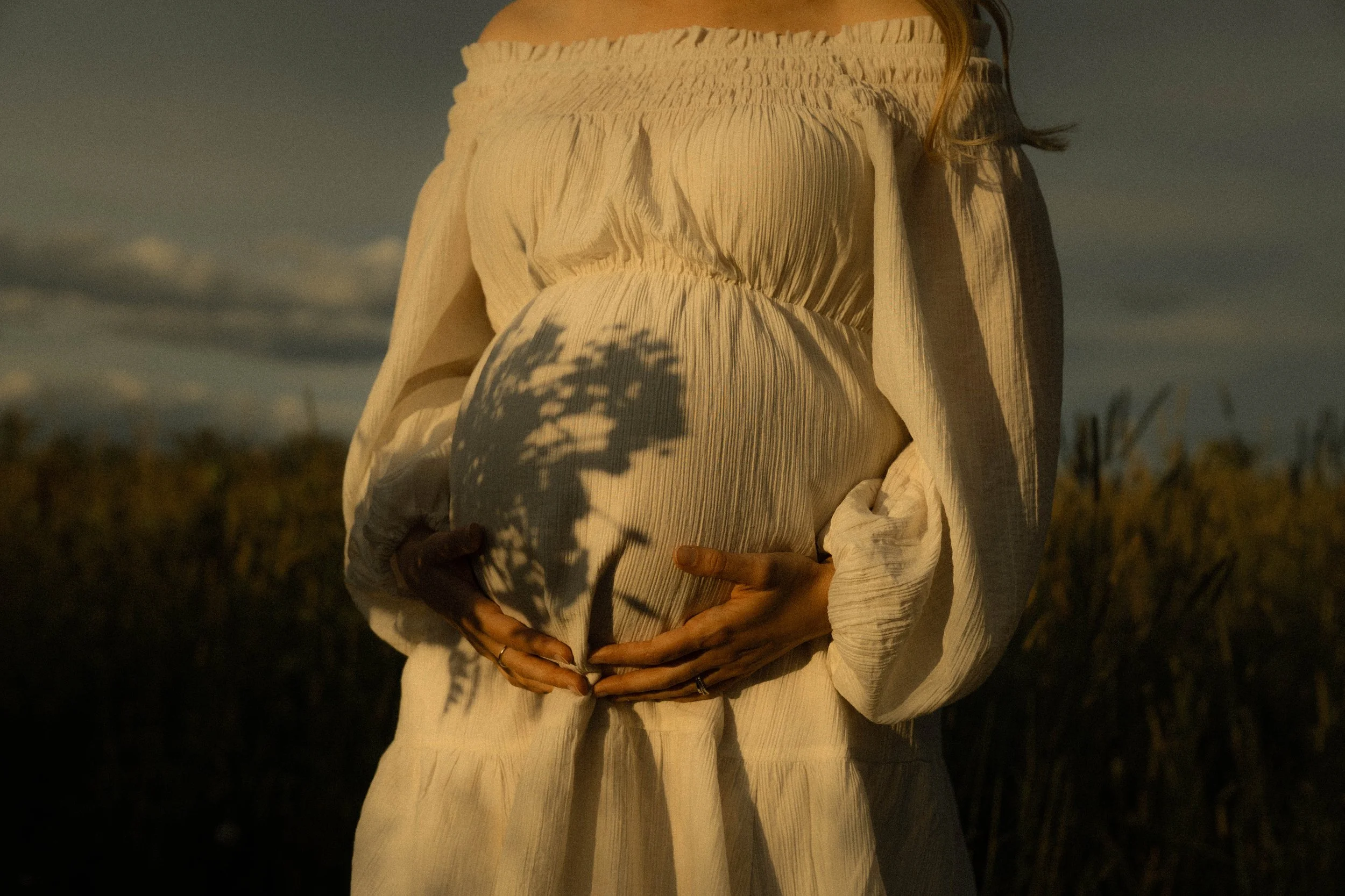 Personne portant une robe beige avec une texture plissée, tenant un objet mystérieux dans la campagne lors du coucher de soleil.