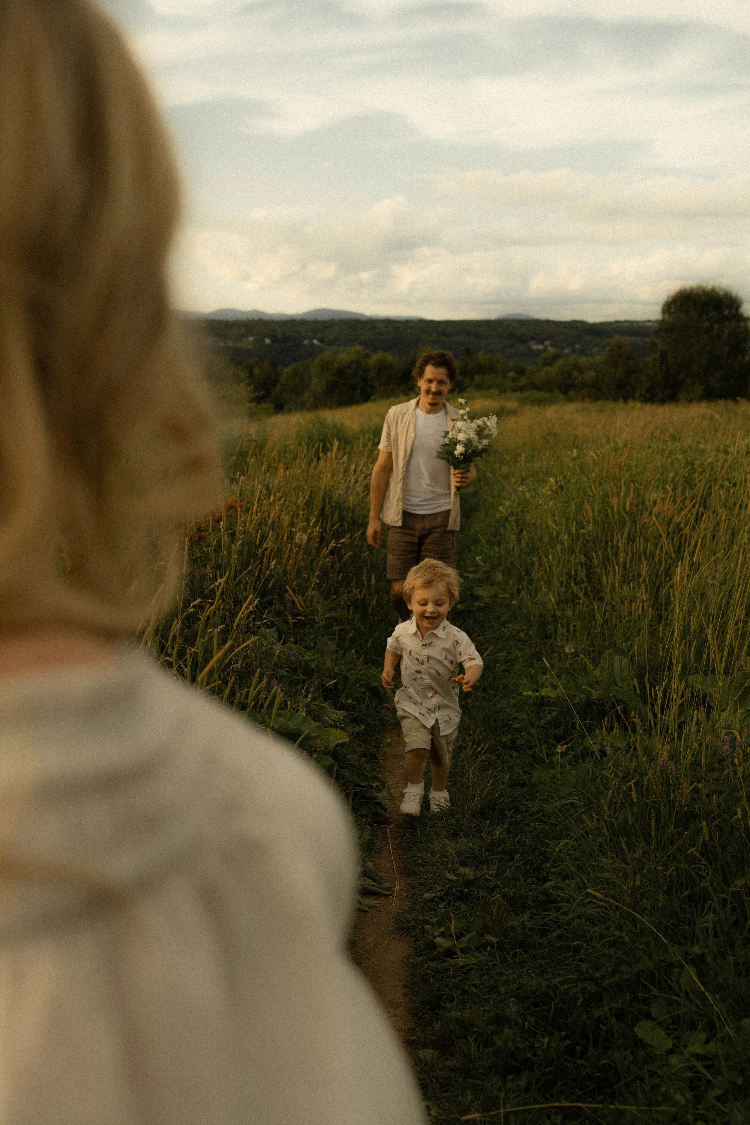 Un homme tient un bouquet de fleurs et marche dans un champ, suivi de deux enfants, dans un paysage rural avec des collines en arrière-plan, ciel nuageux.