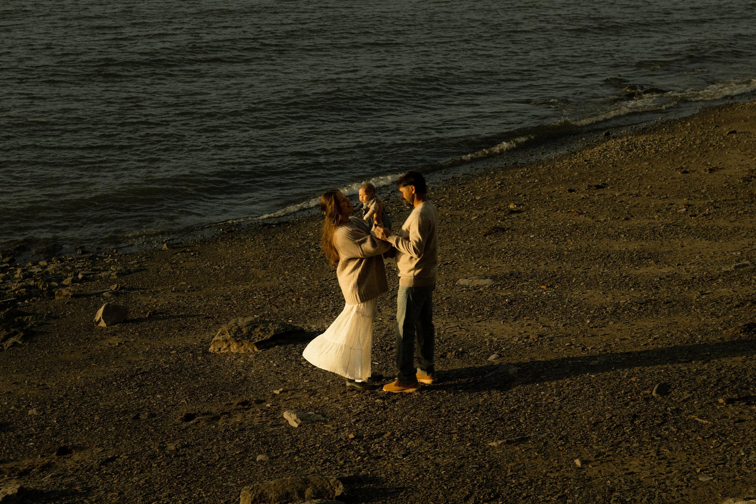 Un couple en tenue de mariage échange des vœux sur une plage au coucher du soleil, avec un bébé entre eux.