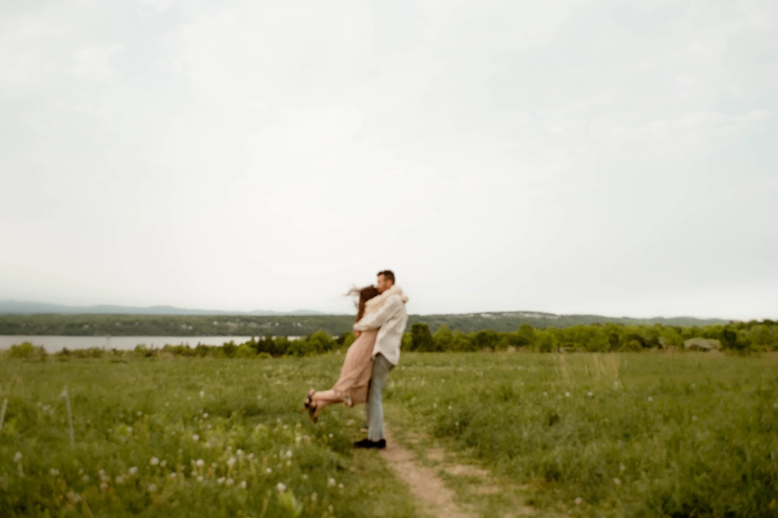 Un couple debout dans un champ d'herbes vertes, s'enlaçant, avec un paysage de nature et un lac au fond, sous un ciel légèrement nuageux.