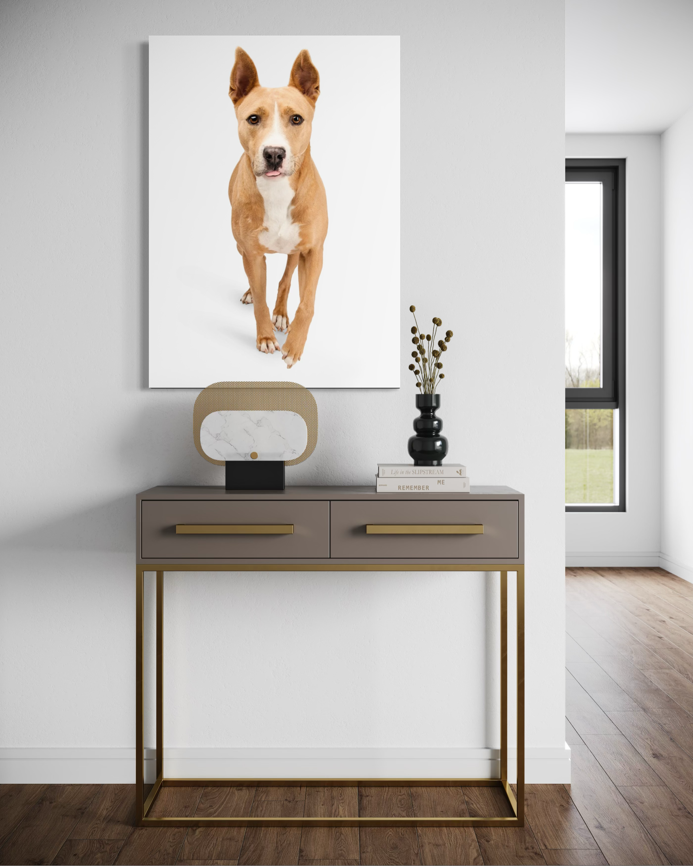 A large framed photo of a tan and white dog hanging on a white wall above a sleek console table with gold accents. The table has a black vase with dried flowers, a small modern lamp, and a few books.