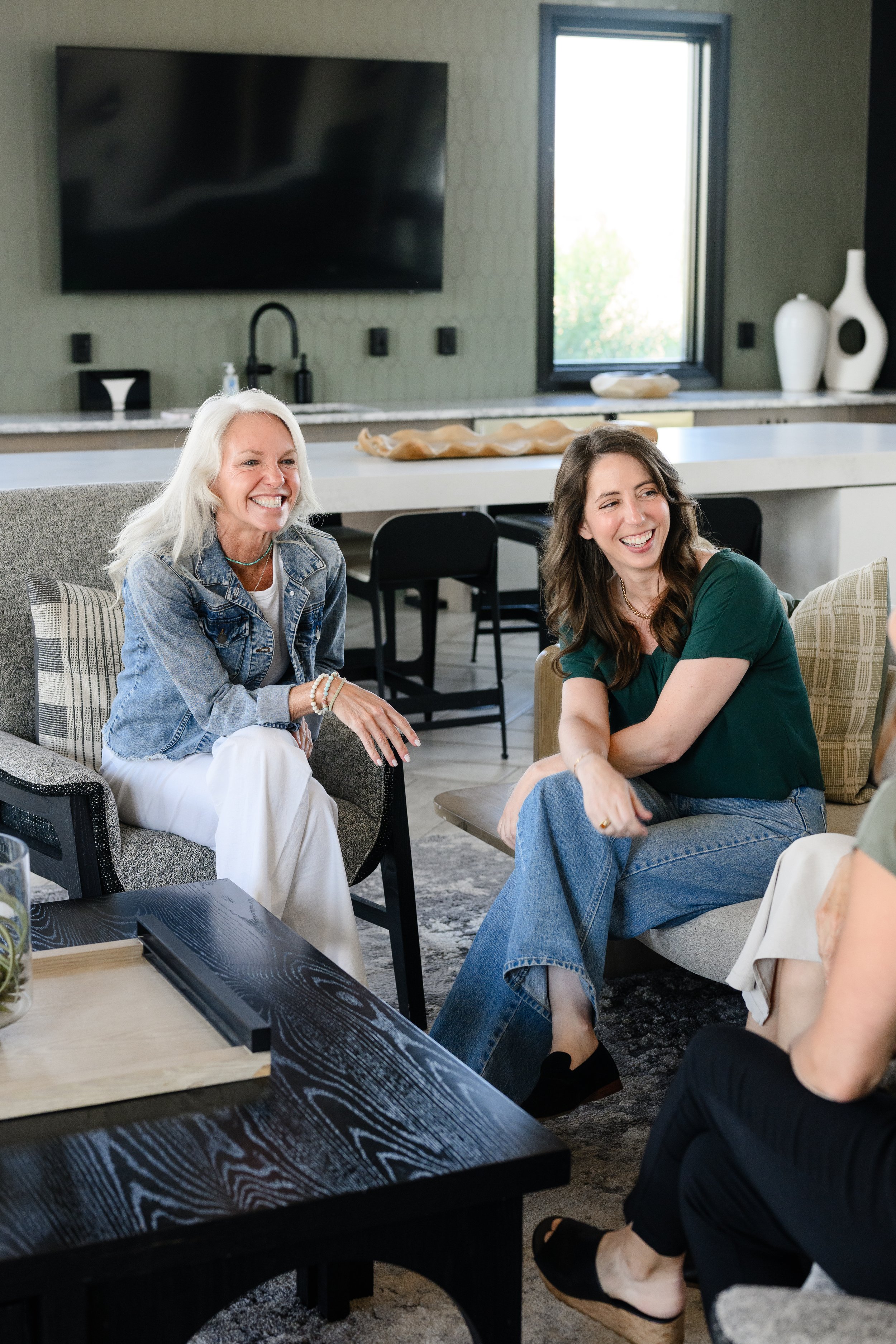 Two women sitting and smiling in a modern living room with a kitchen in the background, including a large TV above the sink, a window with natural light, and decorative vases on the counter.