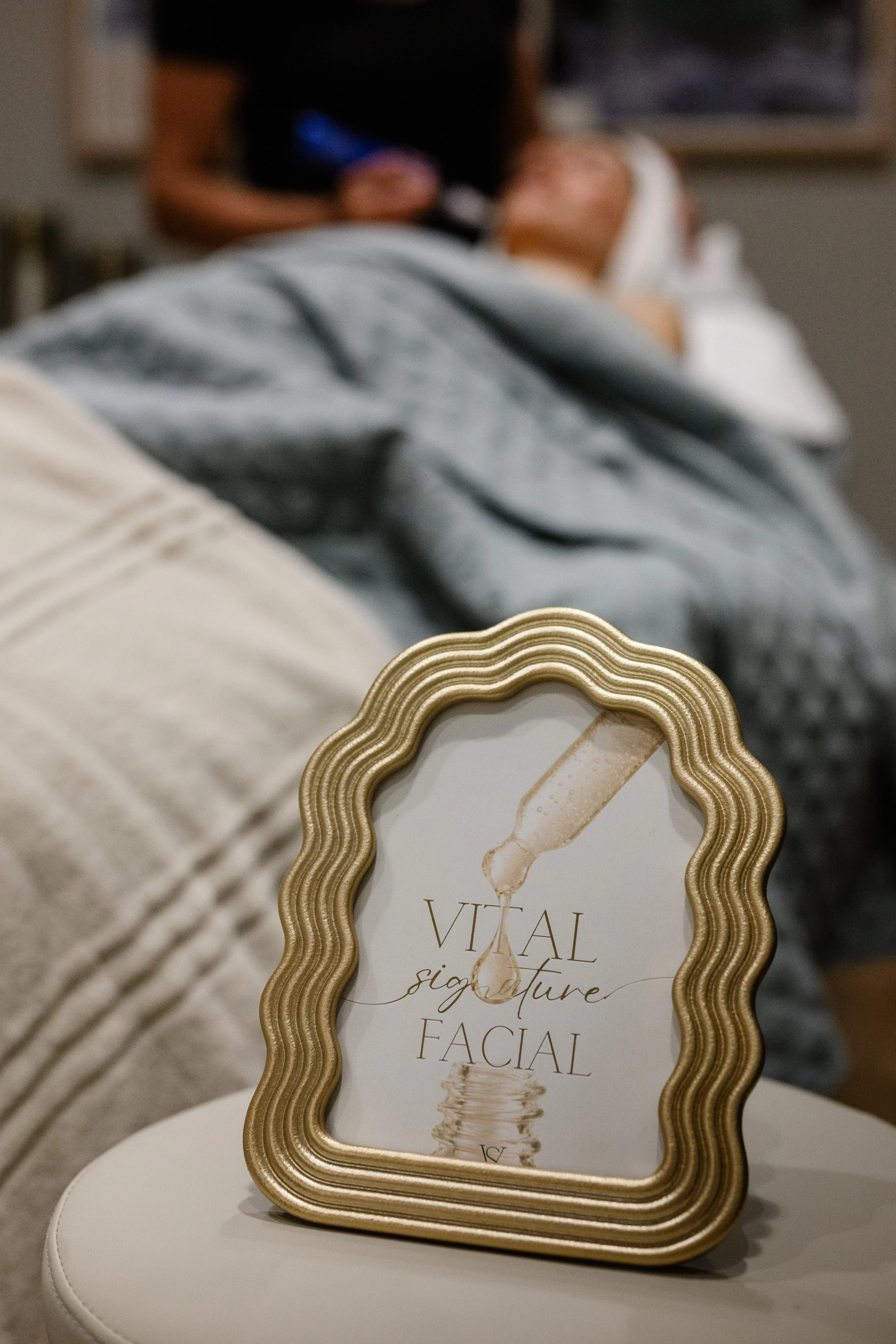 A decorative, gold framed mirror or sign with the text "VITAL signature FACIAL" and a graphic of a dropper above the text. In the background, a person is lying on a treatment bed covered with a cloth, receiving a facial treatment at a spa or skincare clinic.