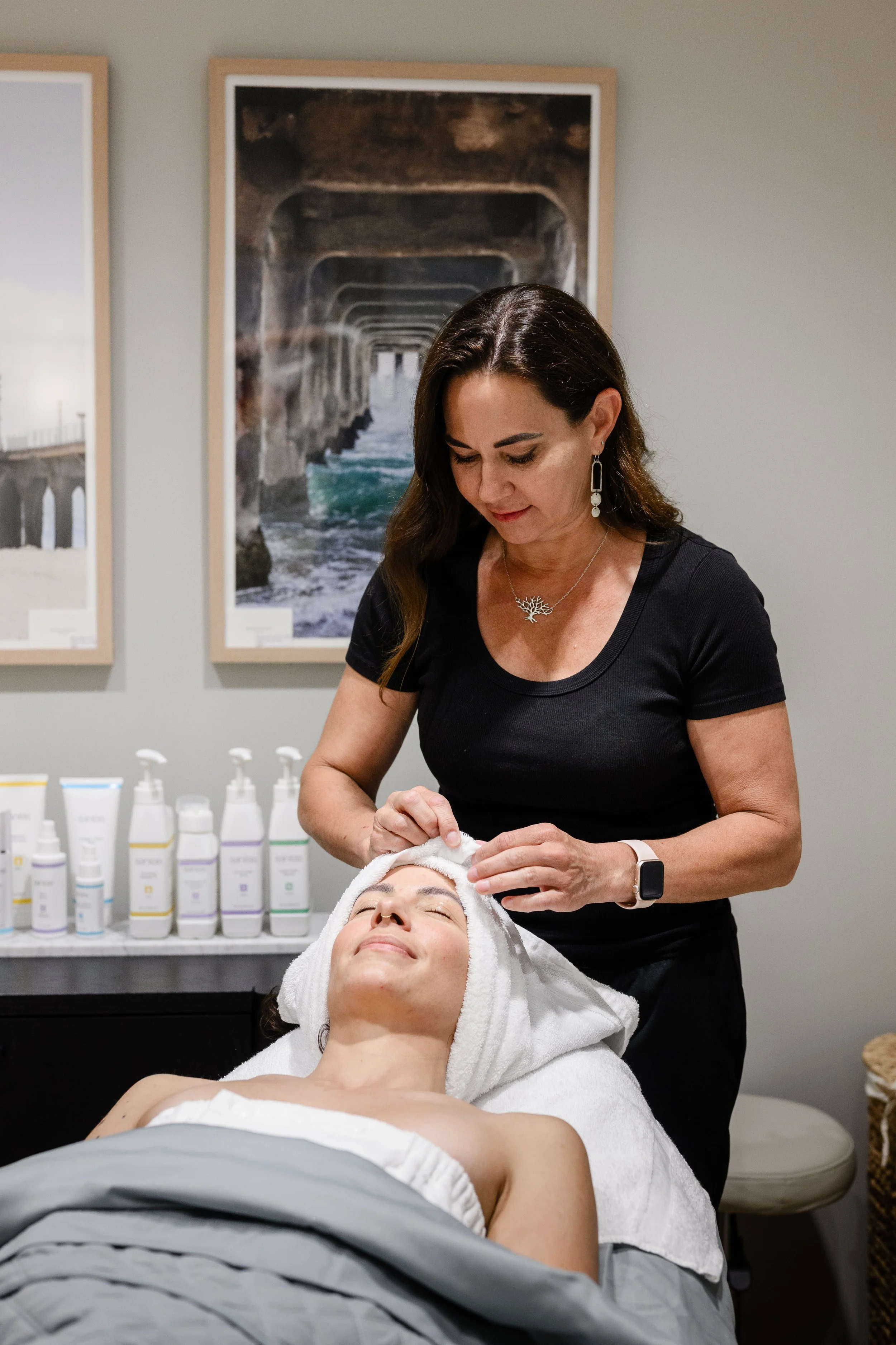 A woman with dark hair wrapping a towel around a client's head to prepare for a facial with Sanitas Nancy brings a steady, caring touch to every visit — helping women feel refreshed, confident, and at home in tprofessional skincare in the background