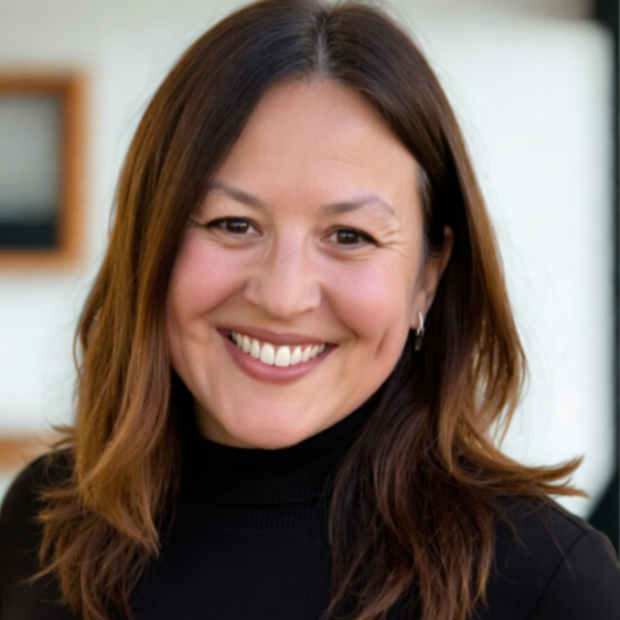 A woman with red hair and tattoos on her arm uses a laptop on her lap, sitting on a teal couch with a white pillow with green and black lines in a living room.