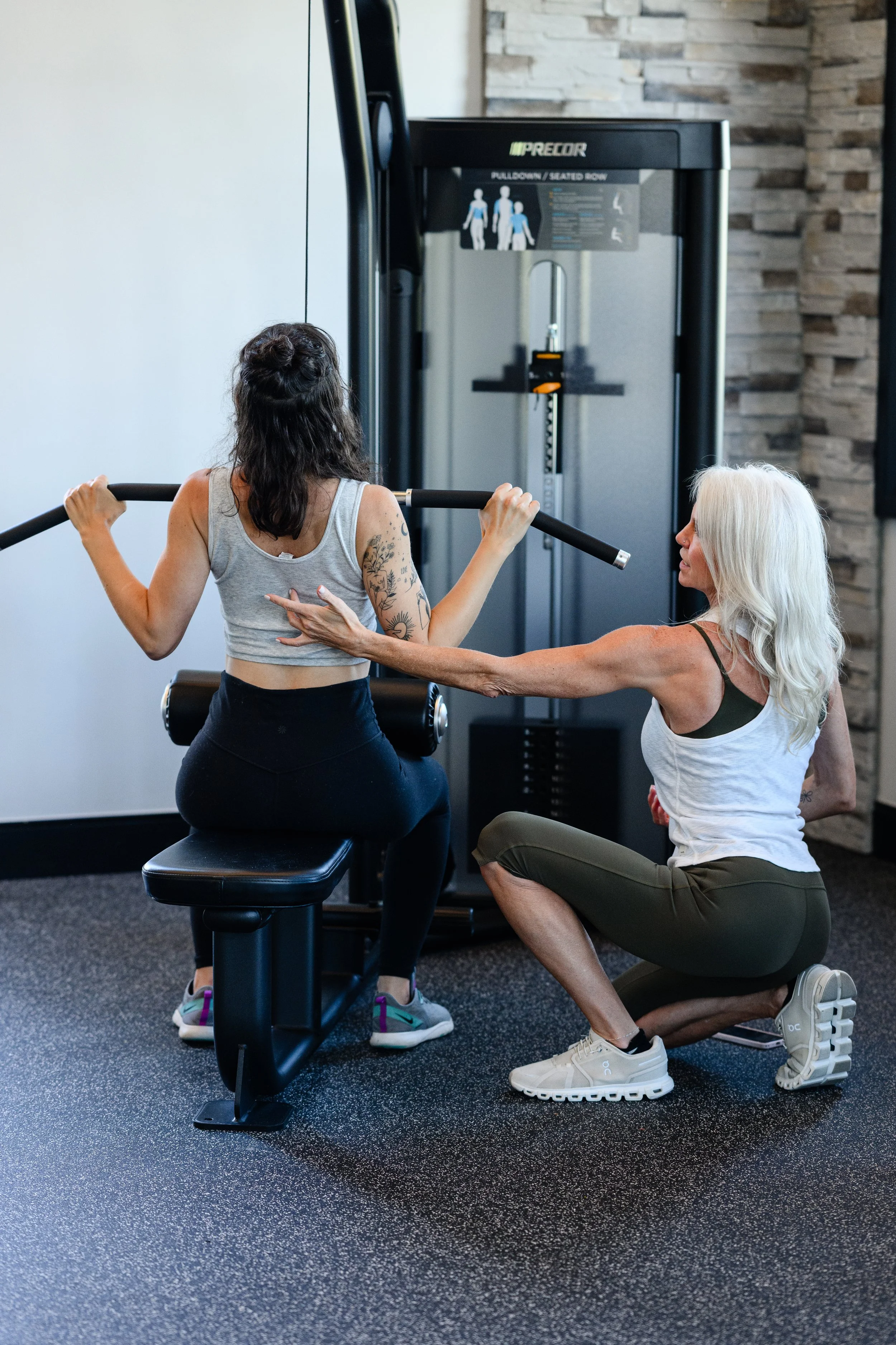 VItal' wellness coach. A woman with long dark hair and tattoos sitting on a workout bench, lifting a barbell, while a trainer with white hair, glasses, and a tank top assists and supports her during a weightlifting session in a gym.