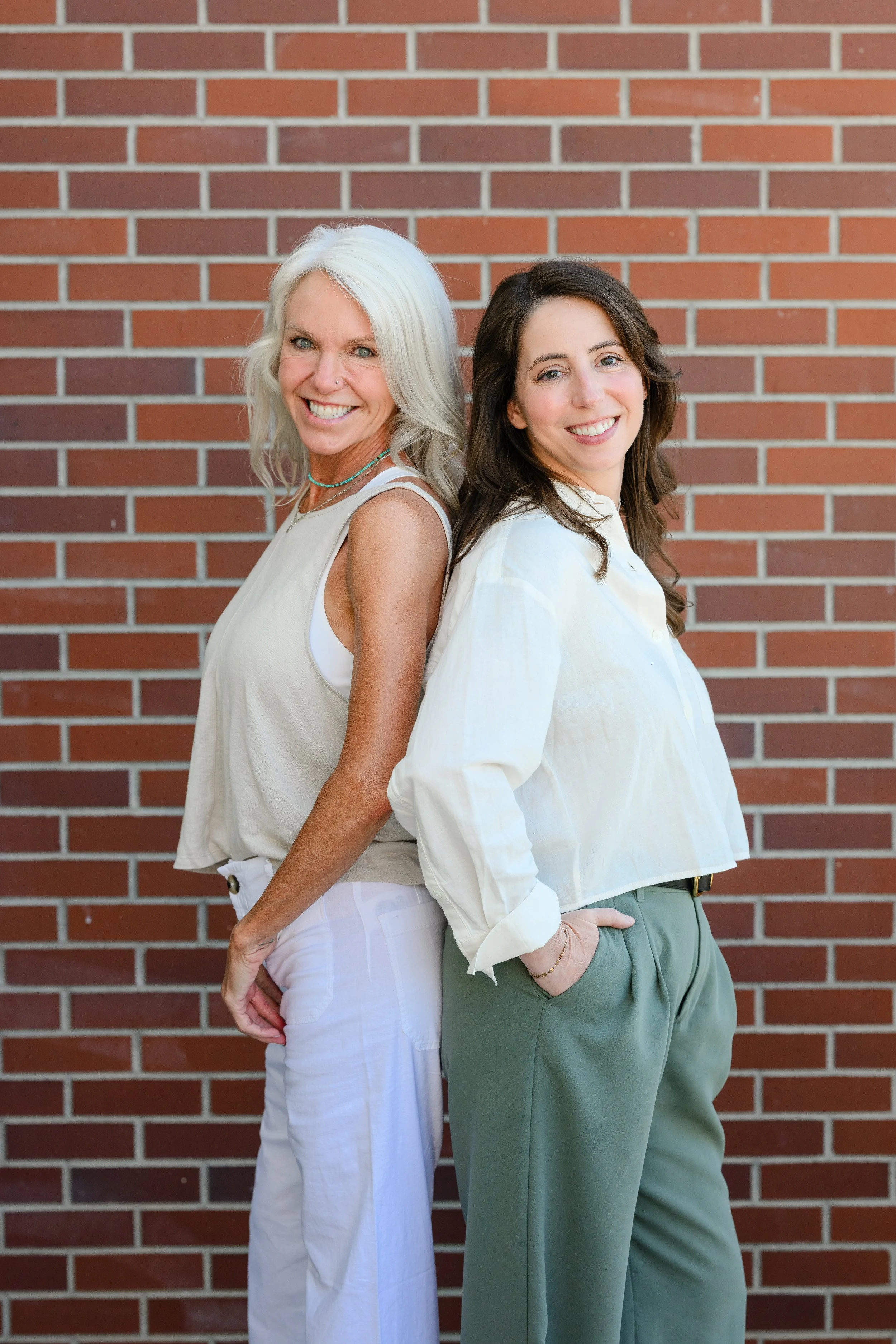 Two women standing back to back smiling, against a brick wall. The woman on the left has long gray hair, wearing a white sleeveless top and white pants. The woman on the right has long brown hair, wearing a white blouse and green pants.