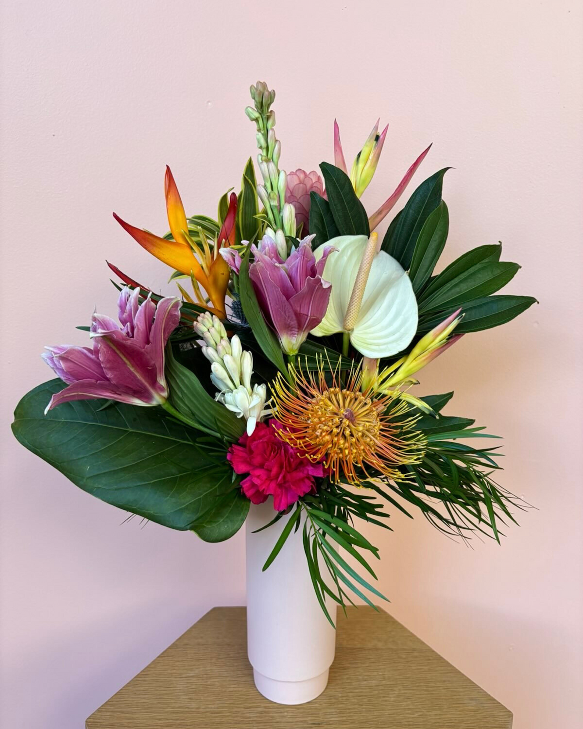 Tropical flower arrangement in a blush pink vase featuring pink lilies, orange pincushion protea, white anthurium, heliconia, and lush greenery, styled by Blūme Ruhm Floral Lounge for same-day delivery in Honolulu, O‘ahu.