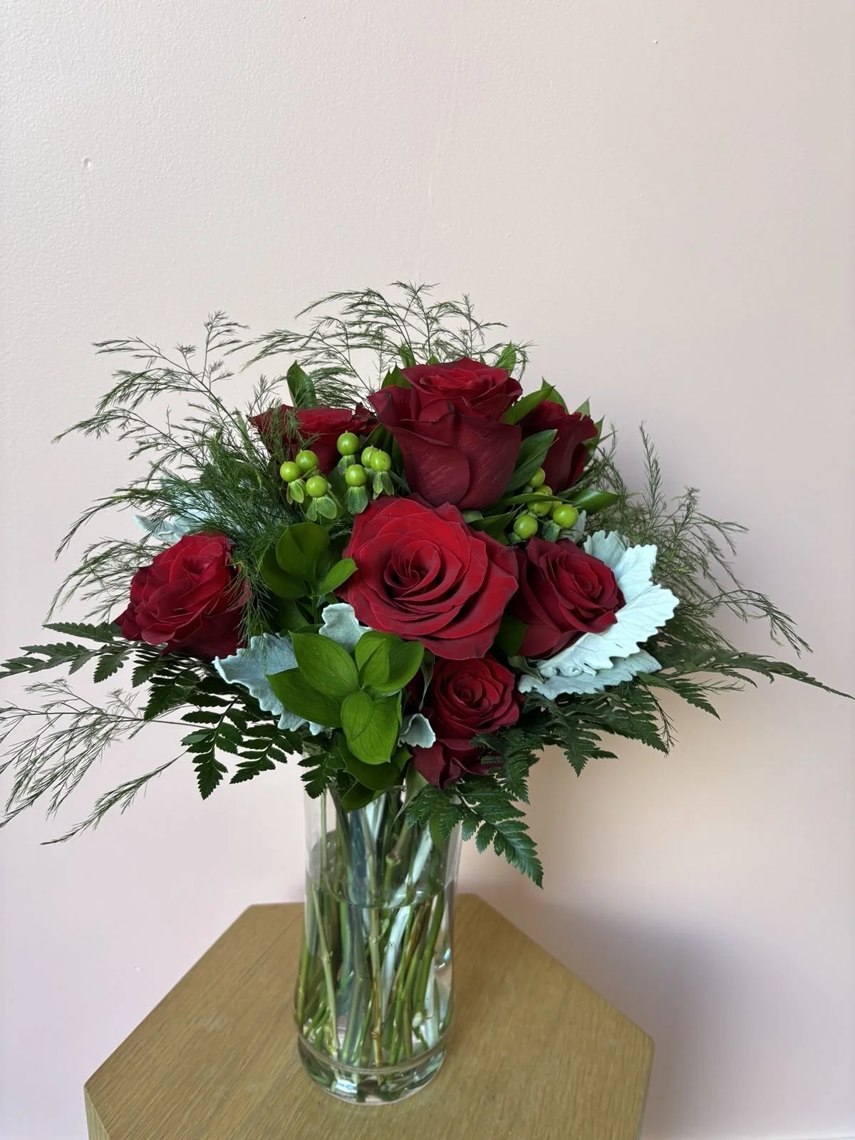 A bouquet of red roses with green berries and greenery in a clear glass vase on a wooden surface.