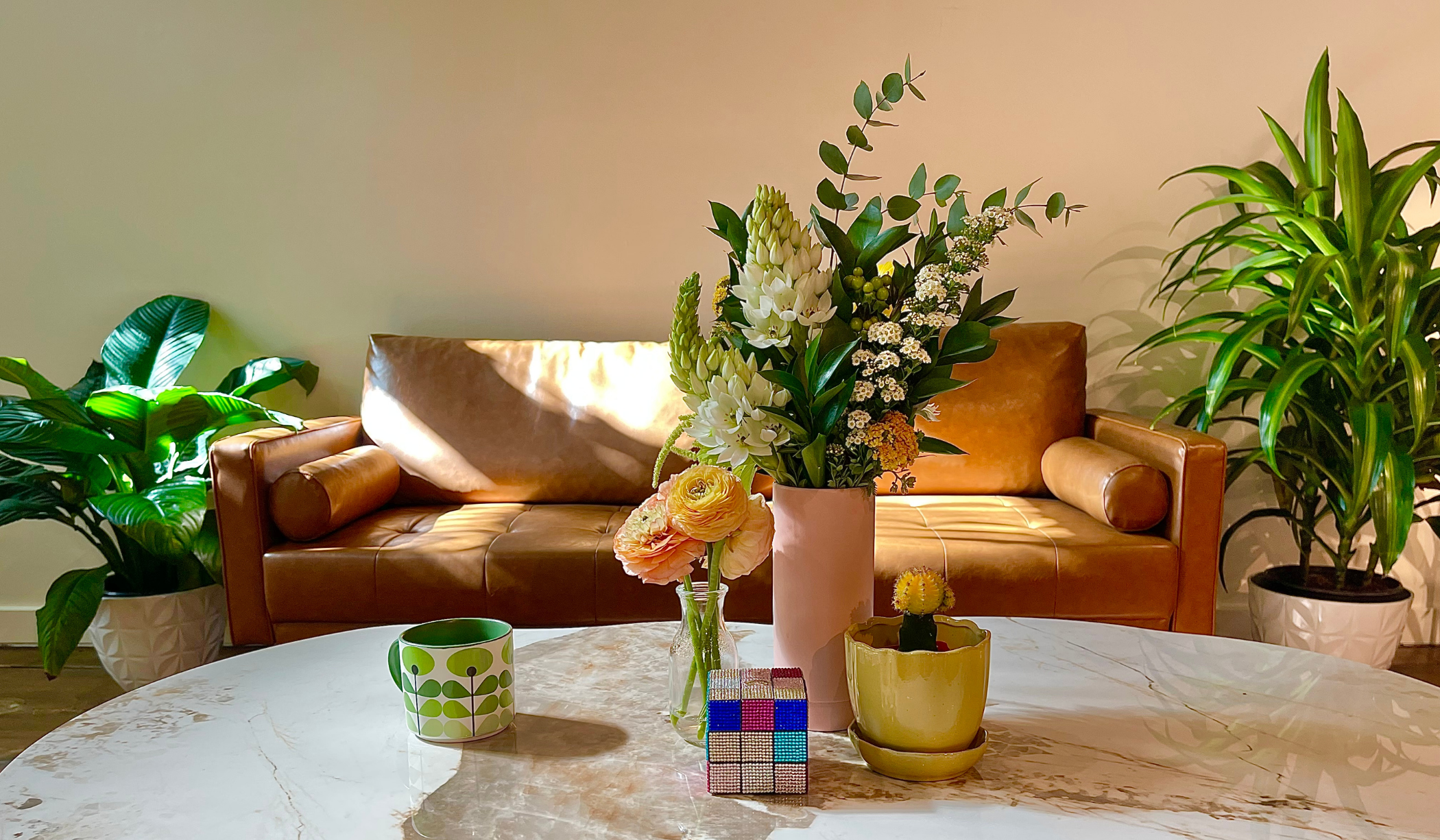 Living room with a white marble coffee table, decorated with a pink vase of white and peach flowers, a small potted cactus, a colorful Rubik's cube, and a green and white patterned mug. In the background, a brown leather sofa flanked by two potted green plants.