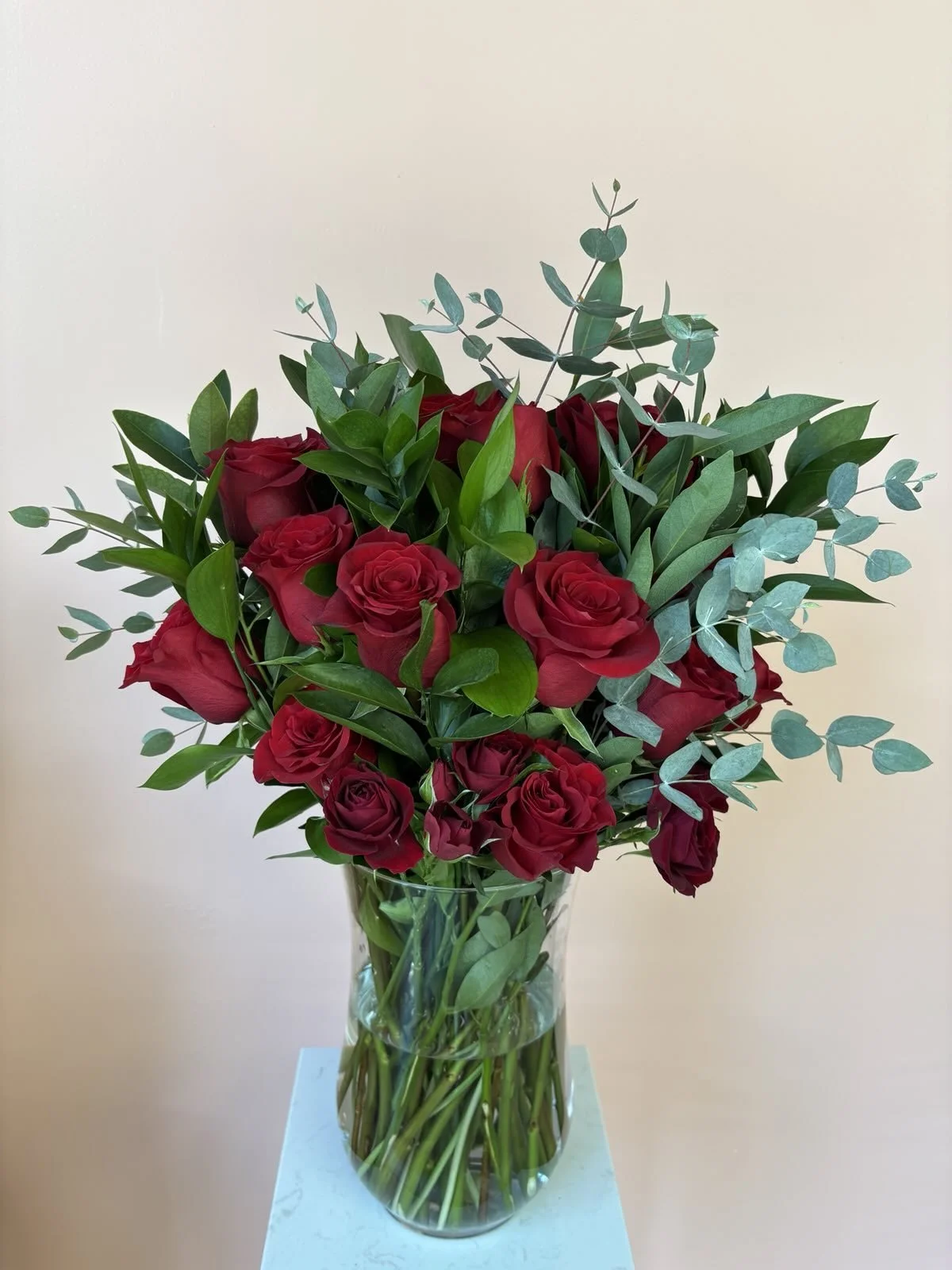 A glass vase filled with red roses and green foliage, placed on a white surface against a plain light background.