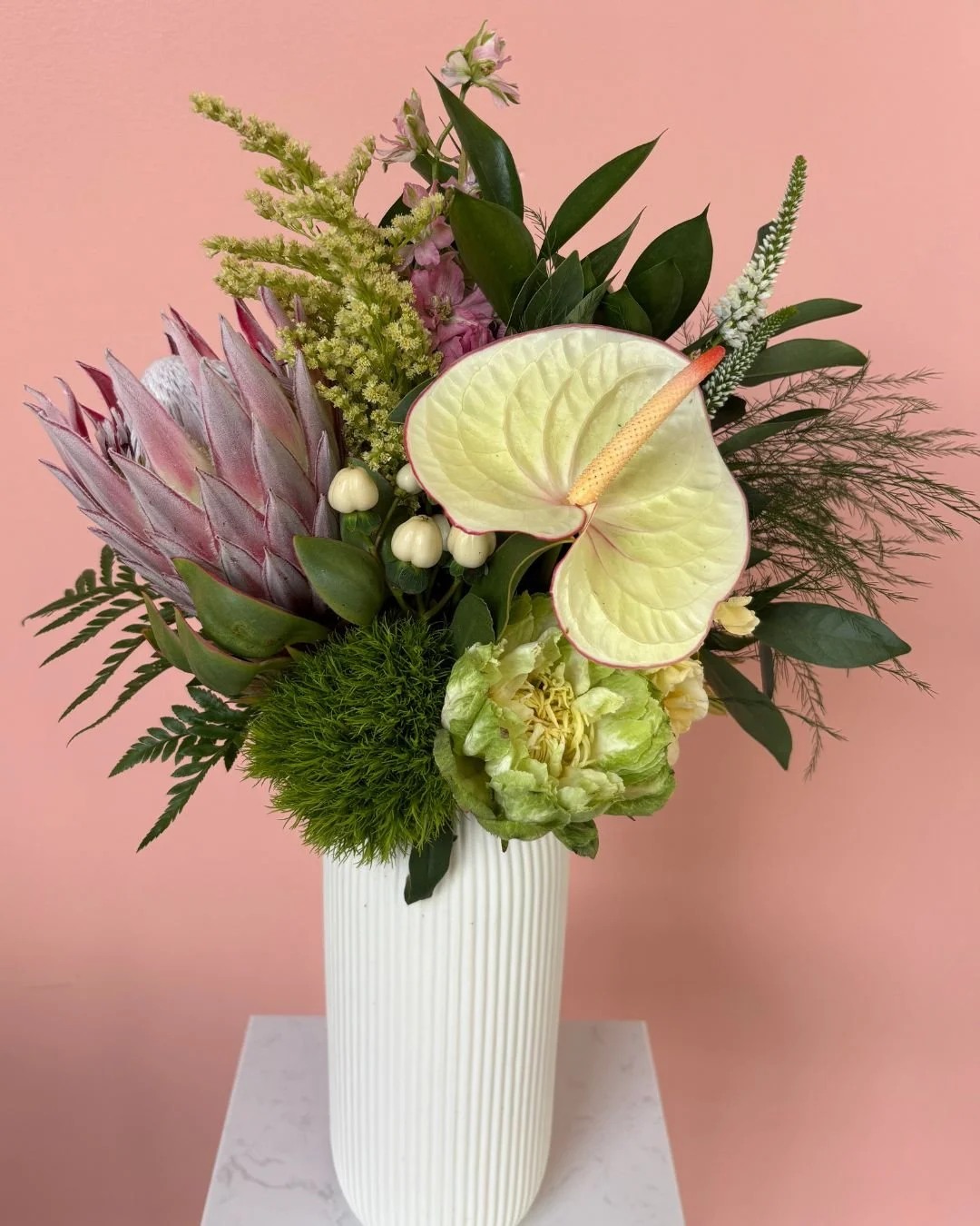 A floral arrangement in a white, ribbed vase against a pink background. The bouquet includes anthuriums, protea, green berries, ferns, and various green leaves.