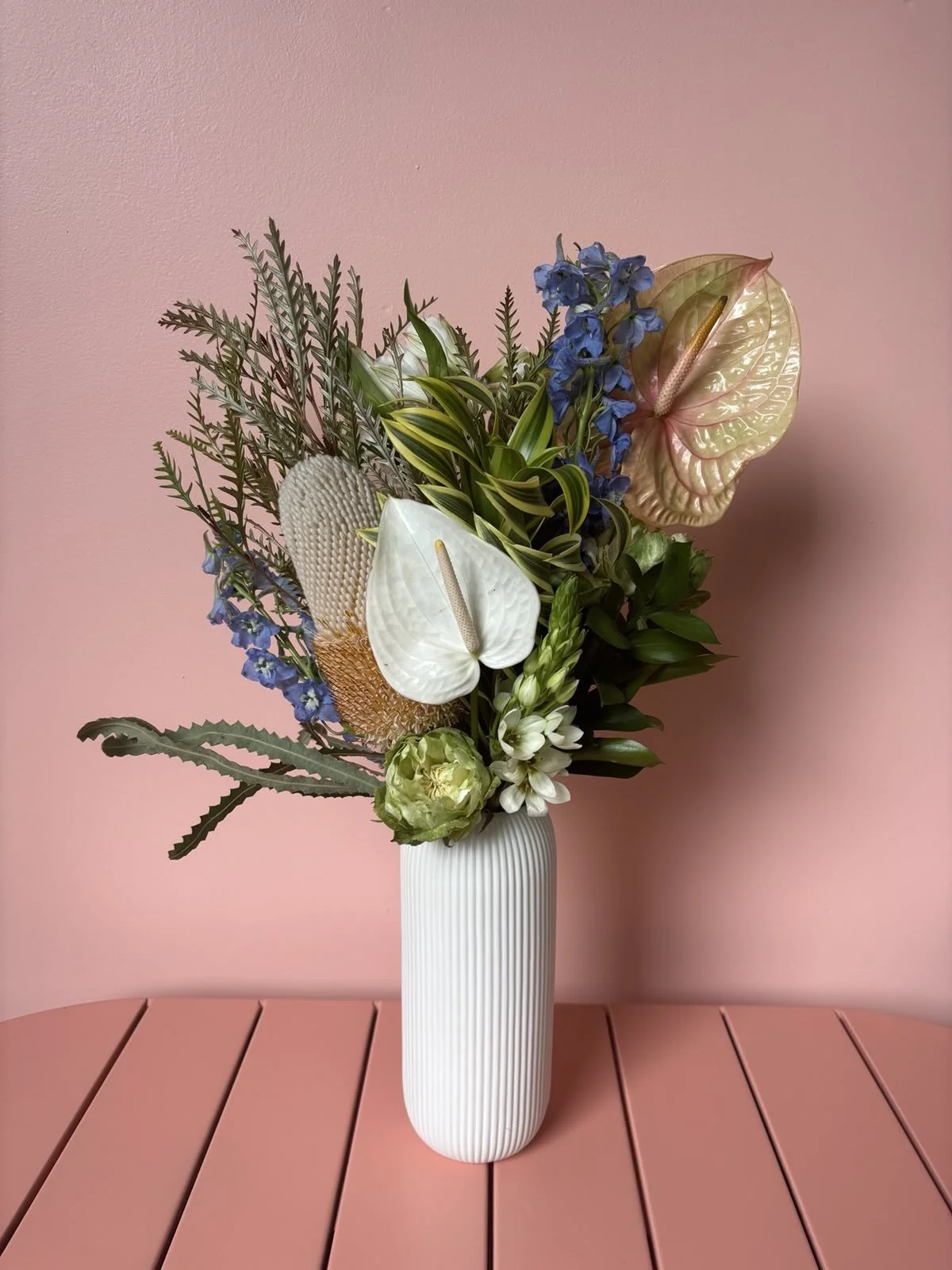 A bouquet of various flowers and greenery in a white ribbed vase on a pink table against a pink background.