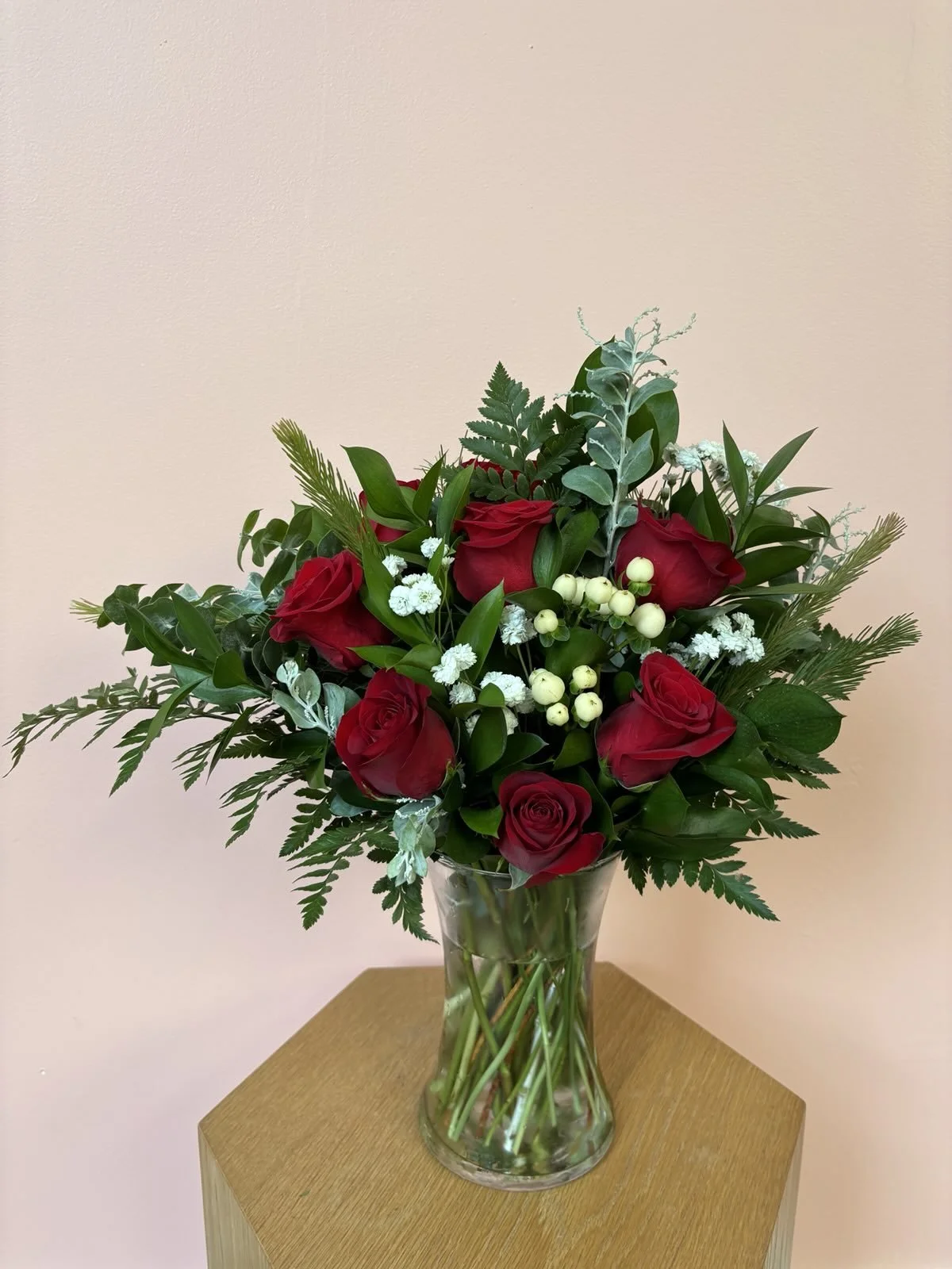 Vase with red roses, white berries, white small flowers, and green foliage on a wooden table.