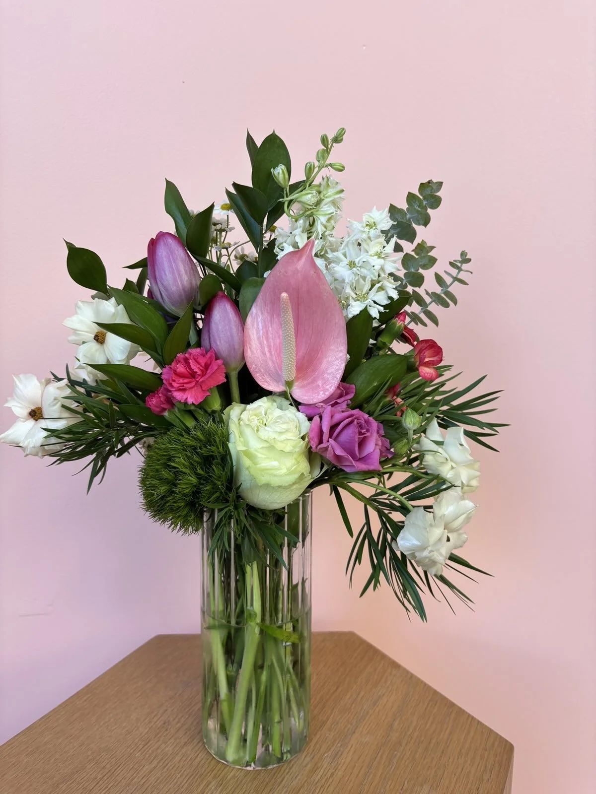 A colorful flower arrangement in a tall glass vase on a wooden table, featuring pink, white, purple, and green flowers with various greenery against a pink wall background.