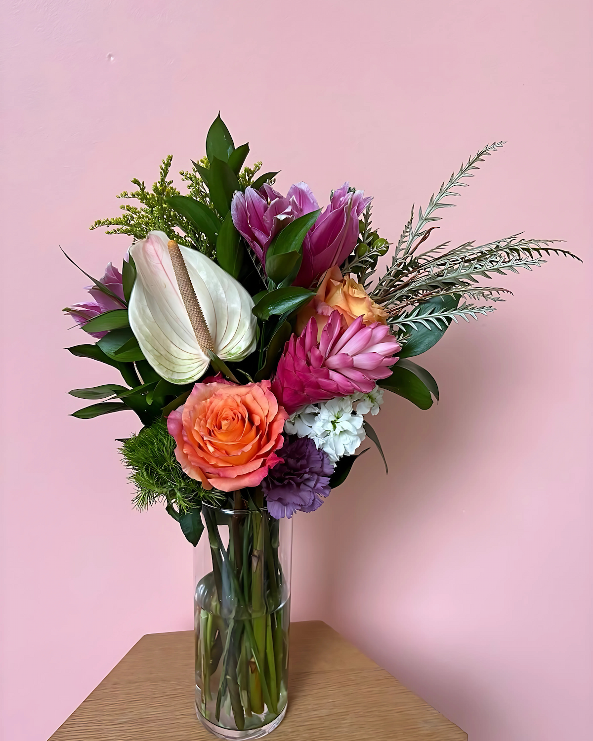 Colorful flower bouquet in a tall glass vase on a wooden surface against a pink wall.