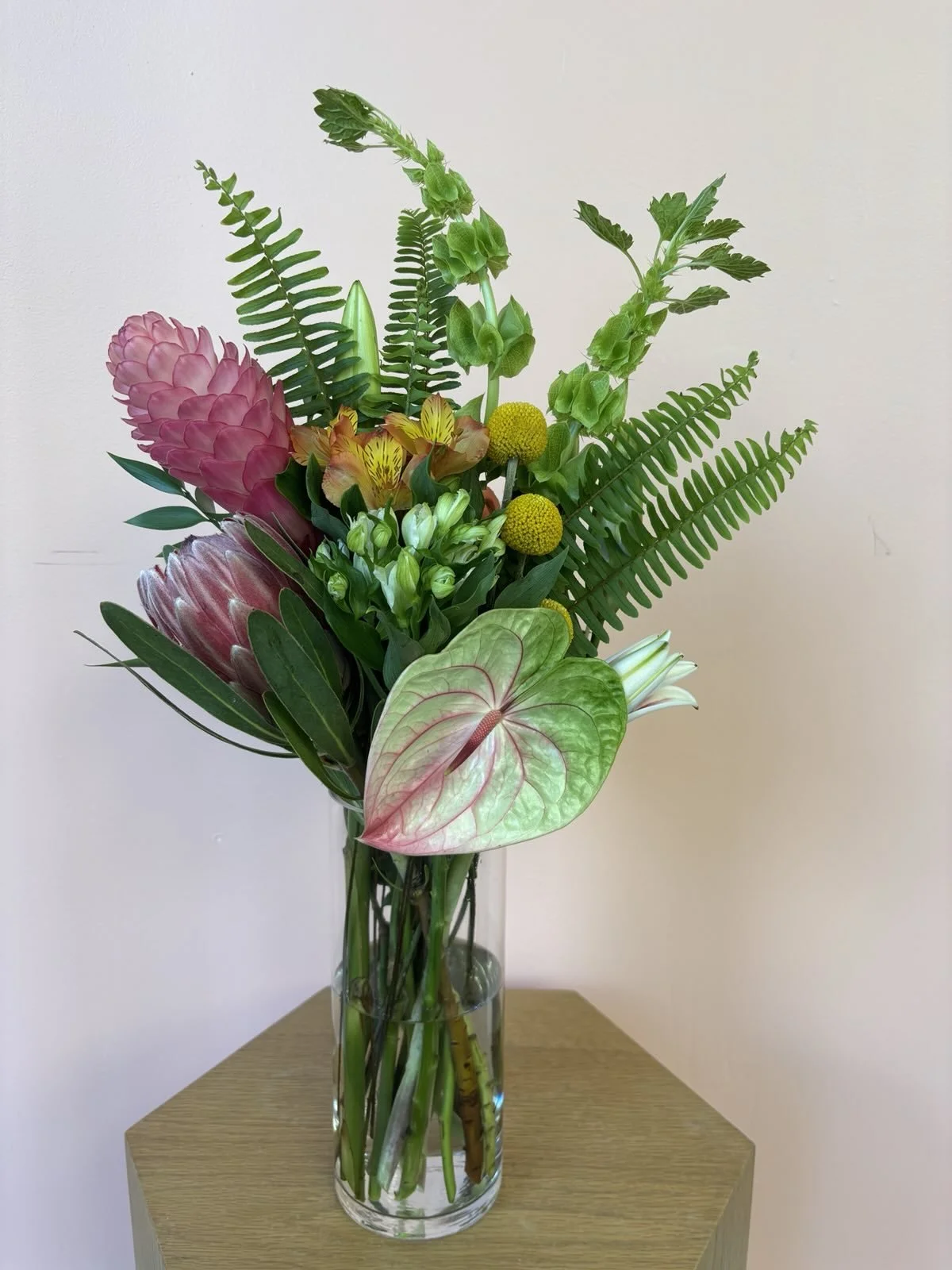 A colorful bouquet of various flowers and green foliage in a clear glass vase on a wooden table against a plain wall.