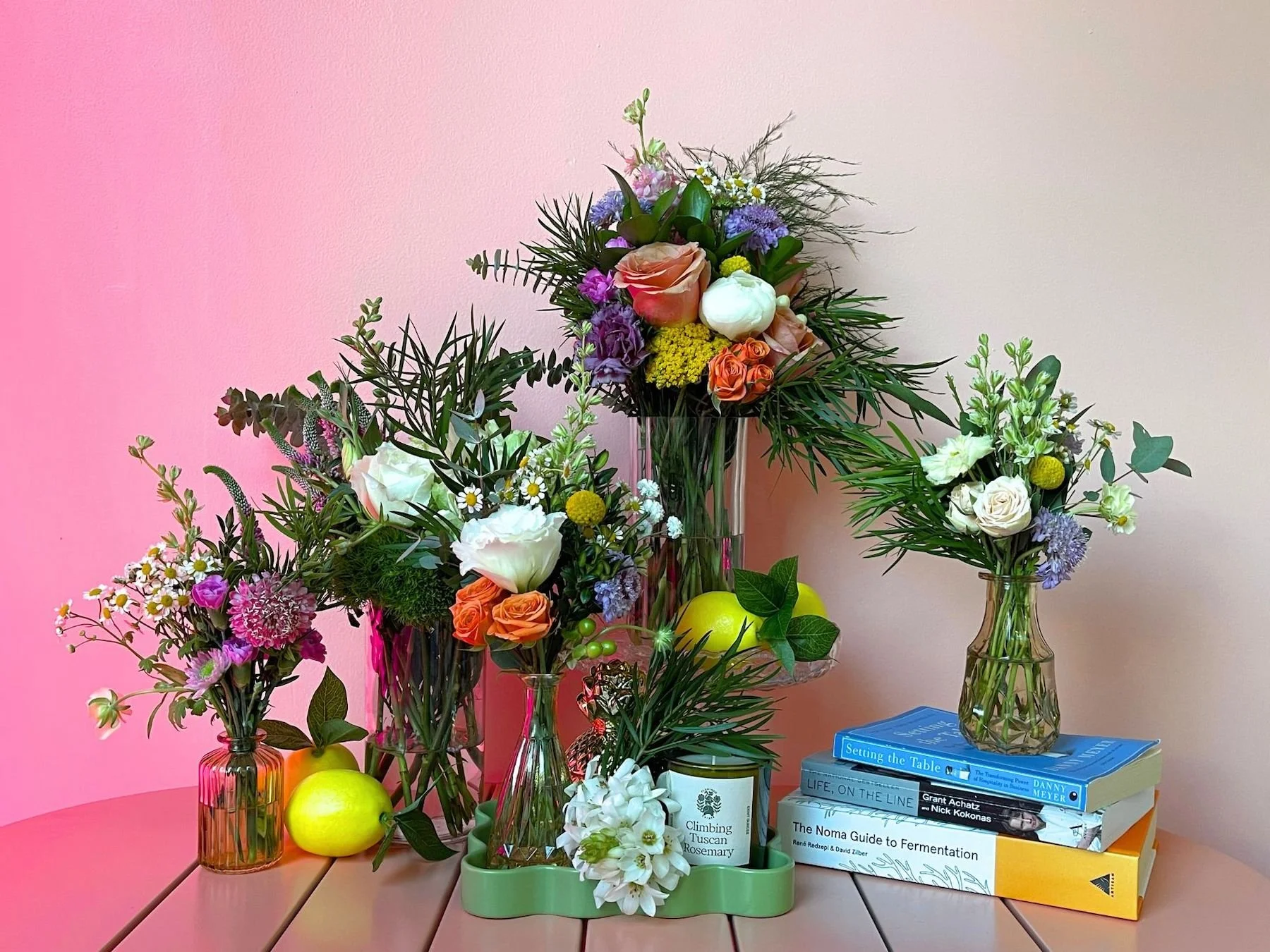 Three vases with assorted colorful flowers and greenery, yellow apples, and a stack of books on a wooden table against a pink and beige wall.