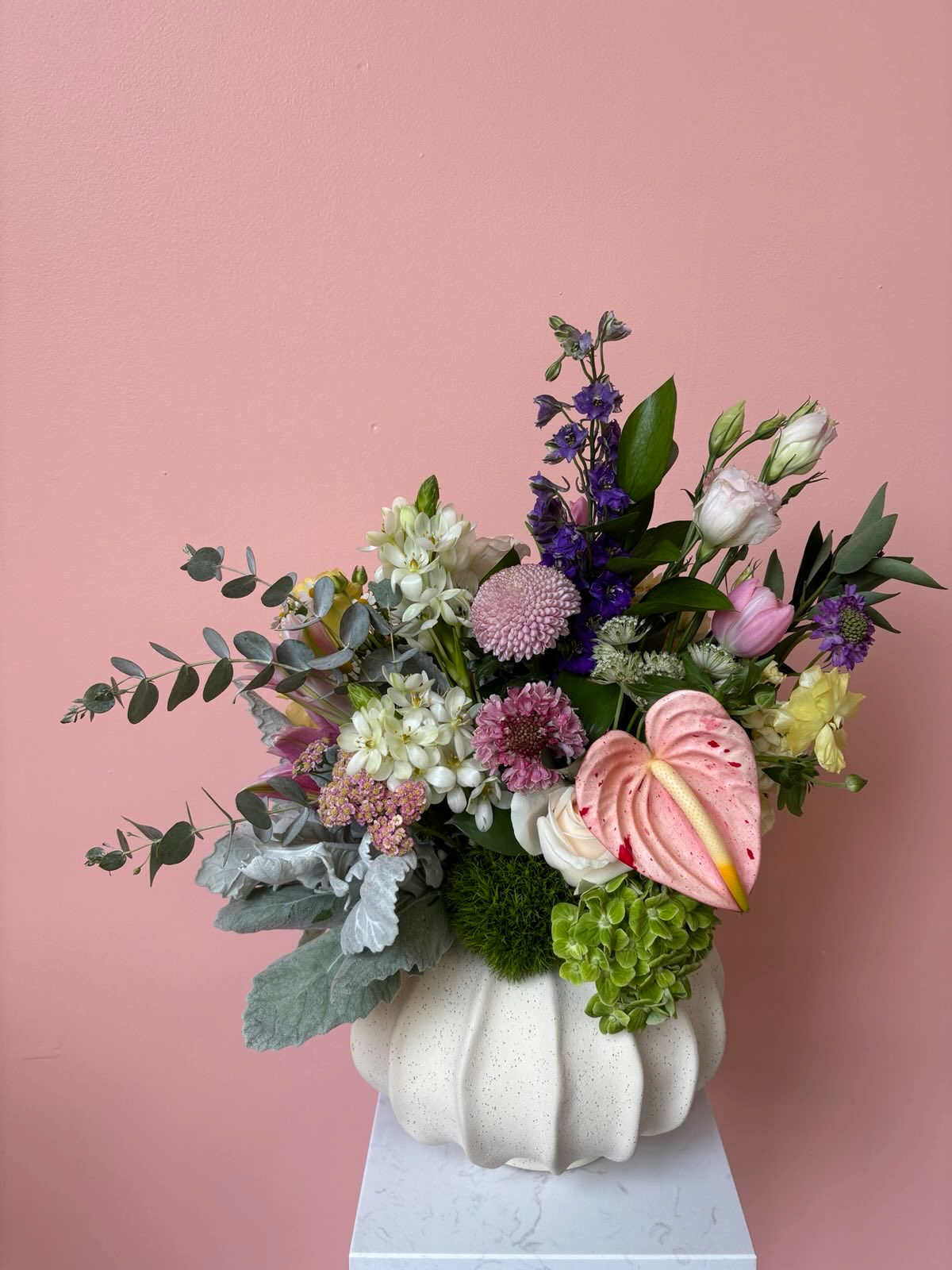 A floral arrangement in a white, textured, pumpkin-shaped vase against a pink wall. The bouquet includes pink, purple, white, and green flowers with greenery and a large pink anthurium.