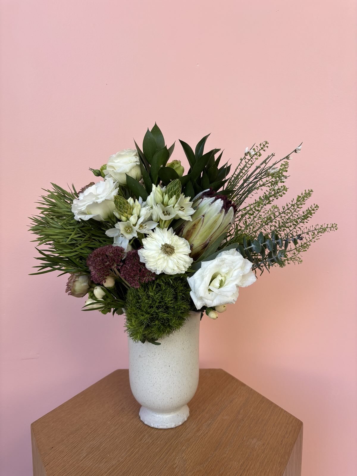 A bouquet of white and green flowers in a white vase on a wooden table against a pink wall.