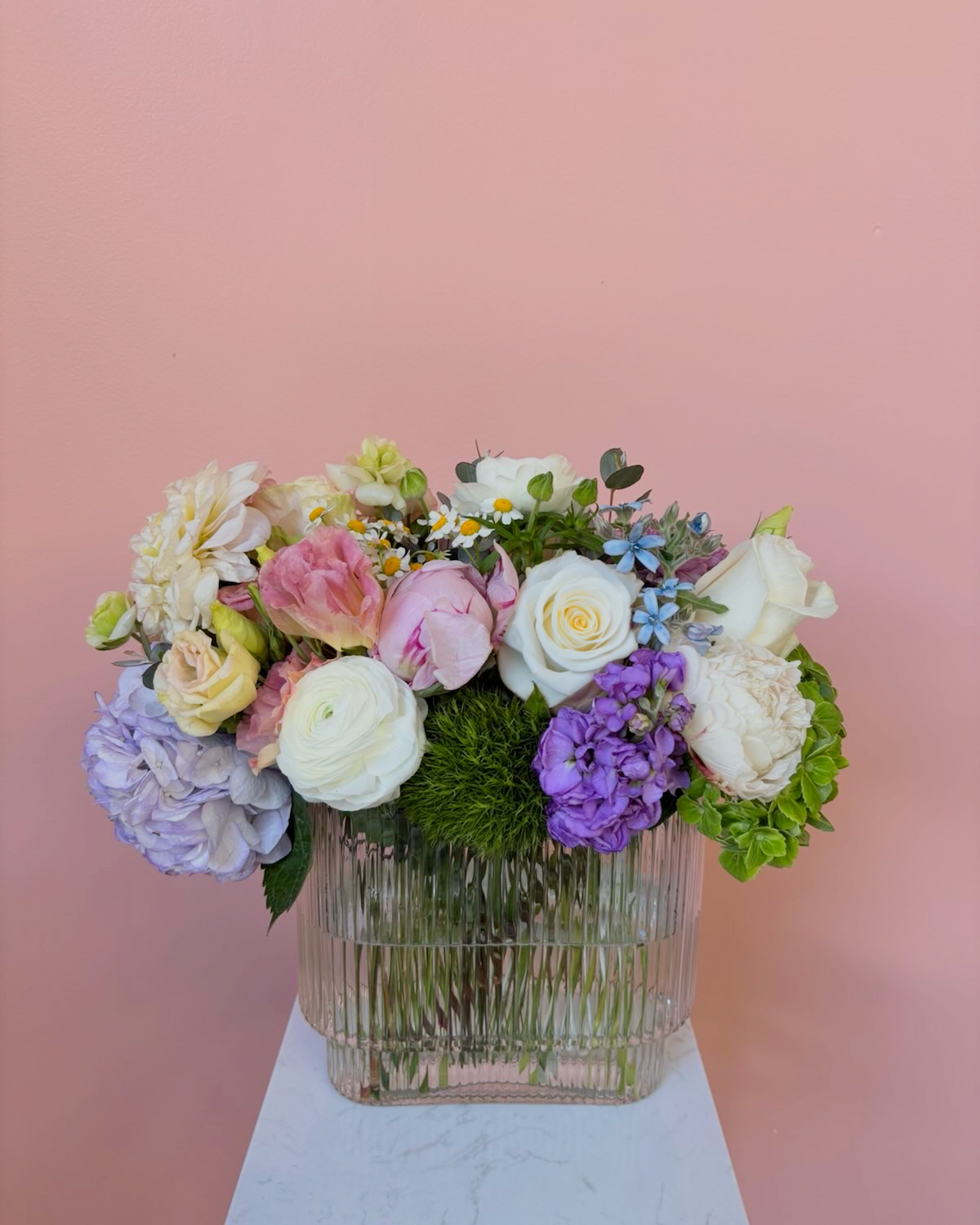 A glass vase filled with a colorful assortment of fresh flowers, including white roses, purple hydrangeas, pink ranunculus, daisies, and greenery, placed on a white surface against a pink background.