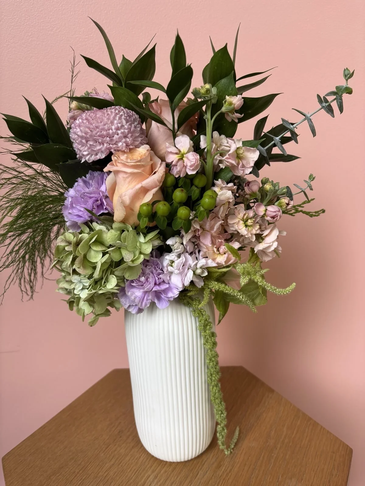 A colorful bouquet of flowers in a white ribbed vase on a wooden table against a pink wall. The bouquet includes pink roses, purple and green hydrangeas, pink and white stock flowers, green berries, and various greenery.