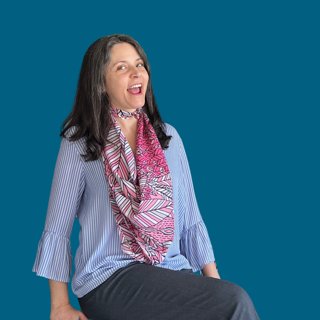 A photograph of a white woman in a seated position. She's wearing a pink scarf against a light blue striped top. Her mouth is open in a big smile. Meet Decluttering Doula, a decluttering expert in the Seattle area.