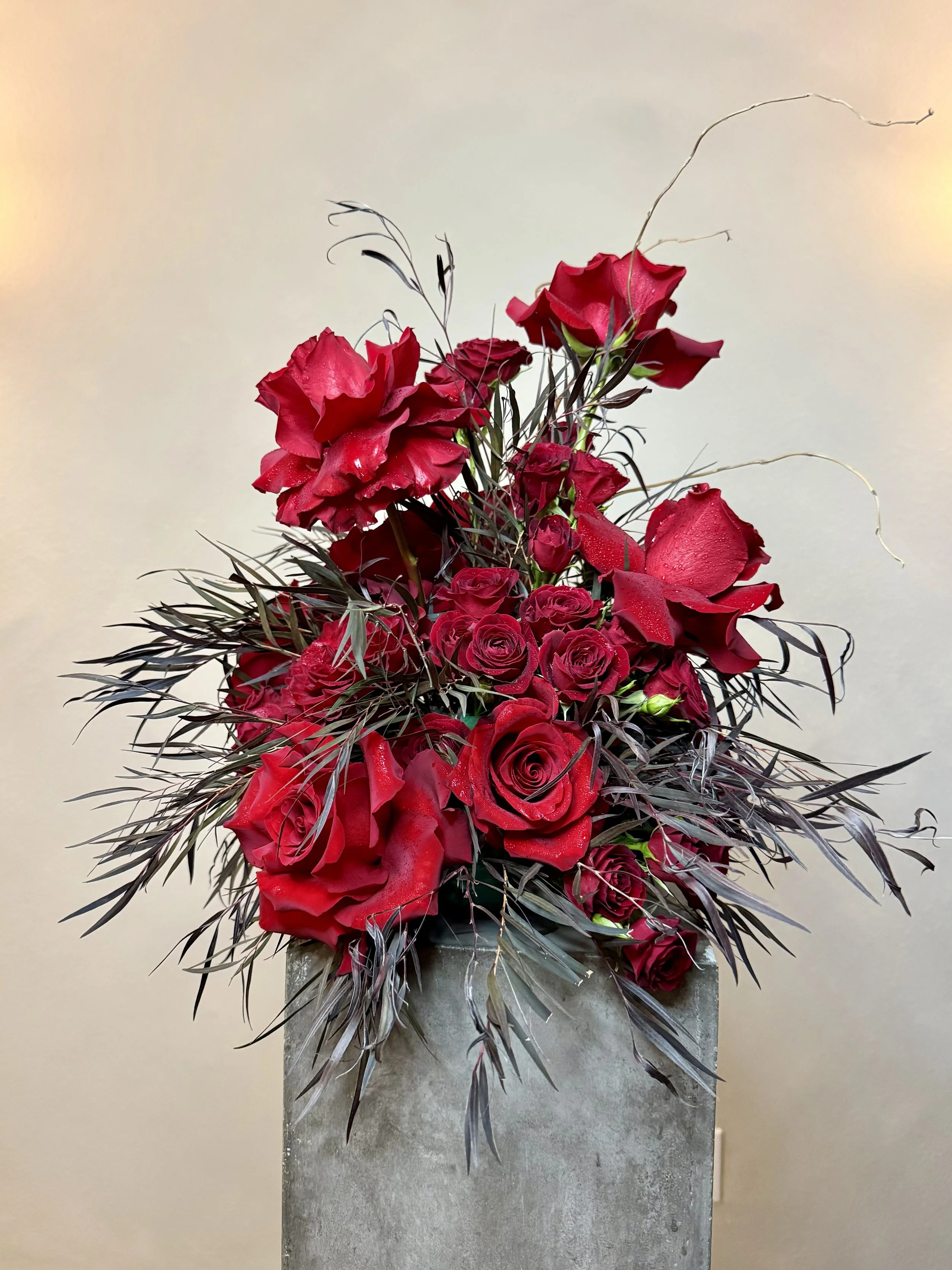 A floral arrangement featuring red roses, red calla lilies, and dark purple foliage in a concrete vase.