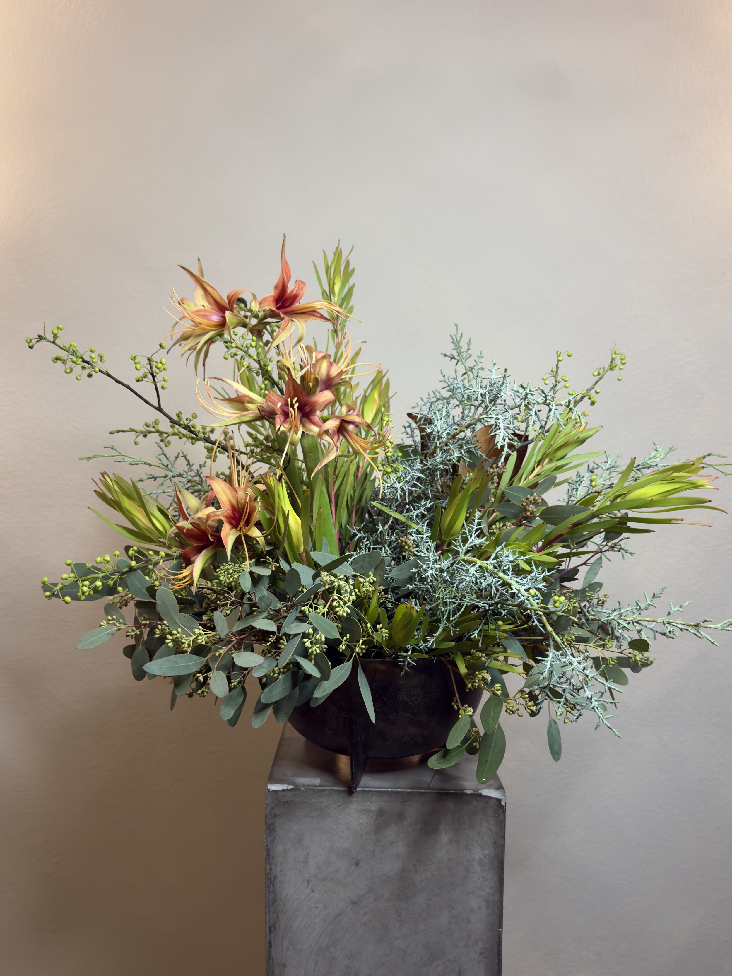A floral arrangement with eucalyptus, orange lilies, and other greenery in a black bowl on a concrete pedestal against a plain beige wall.