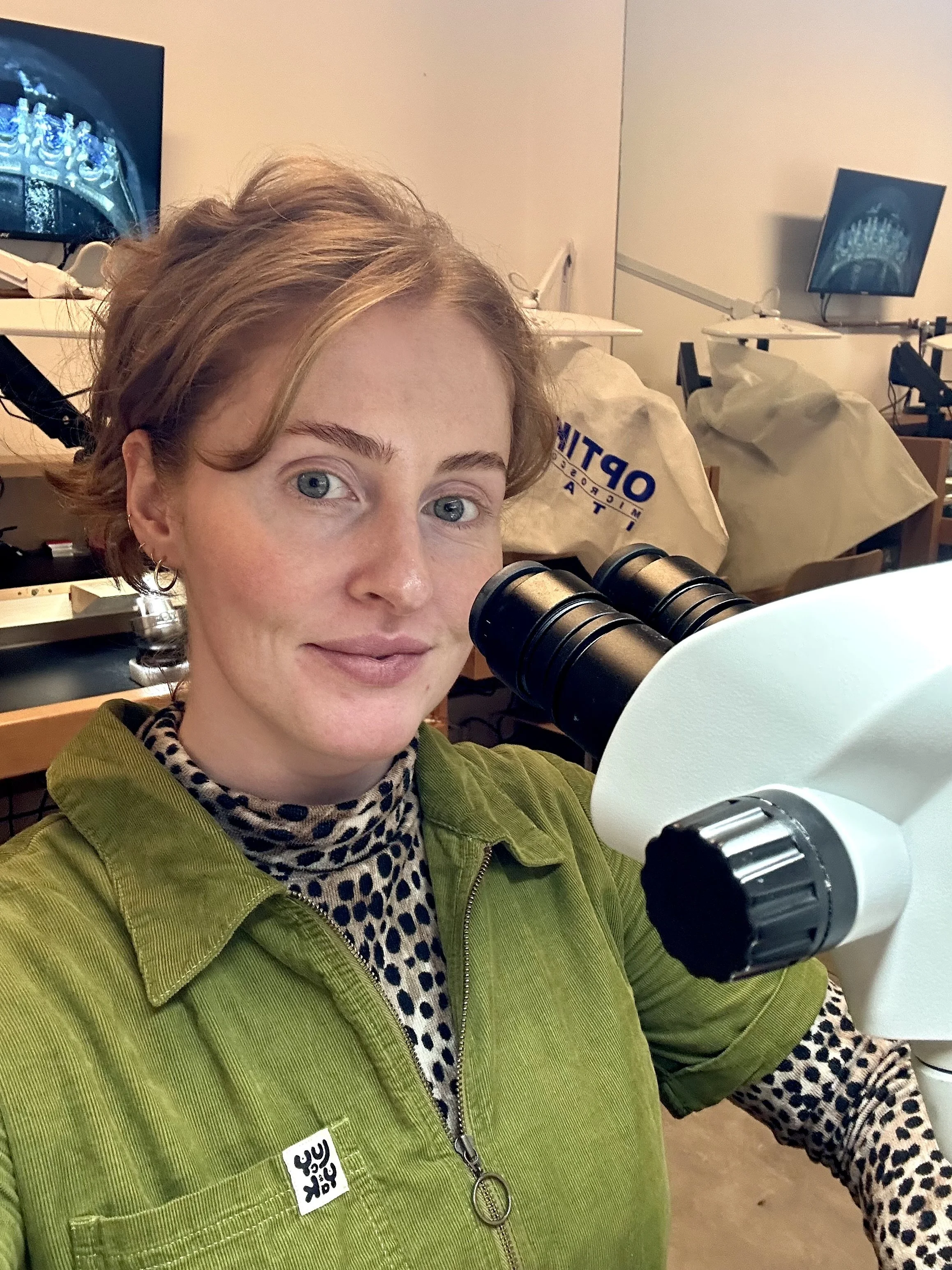 A woman with short wavy hair and blue eyes looking at the camera, using a microscope in a laboratory setting with computer monitors and equipment in the background.