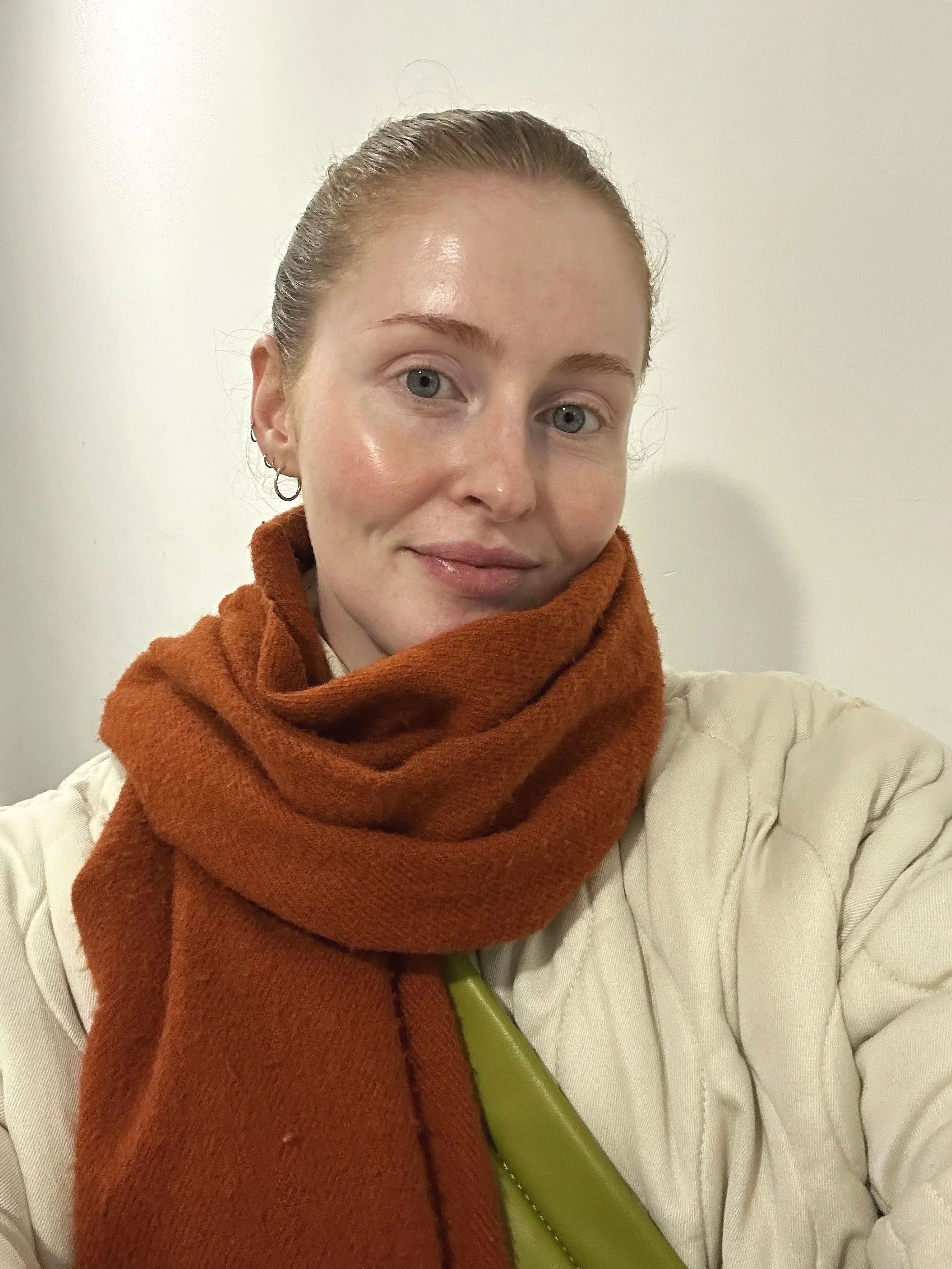 A woman with light skin, red hair pulled back, wearing a beige quilted jacket and an orange scarf, taking a selfie against a plain white wall.