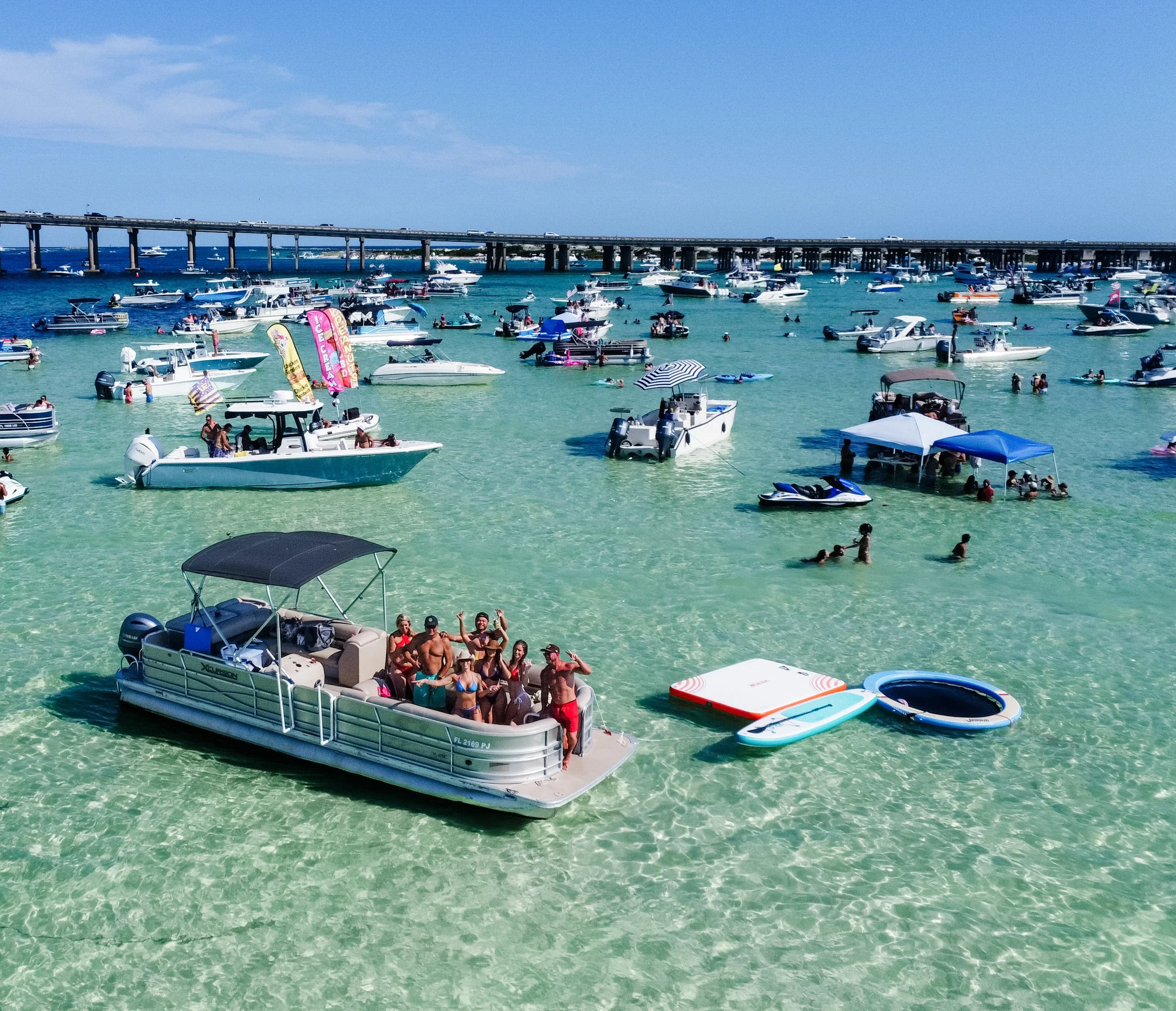 Crab Island Destin Florida with boats anchored in clear turquoise waters, people swimming and lounging, and a bridge in the background under a blue sky.