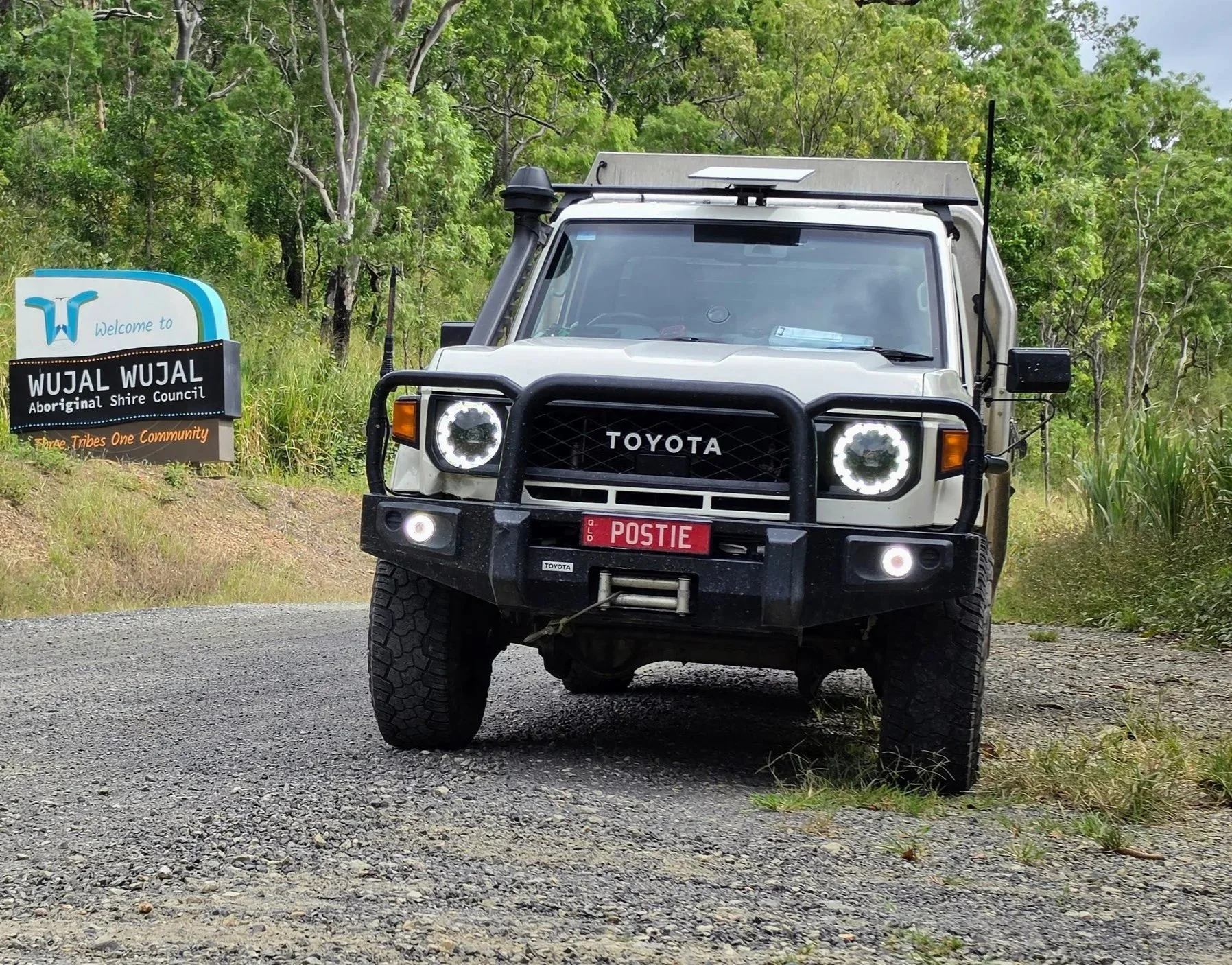 A white Toyota off-road vehicle with a black bumper and large tires parked on a gravel area beside a wooded area. There is a signboard that reads 'WUJAL WUJAL Aboriginal Shire Council' and 'Three Tribes. One Community' on the left side of the image.
