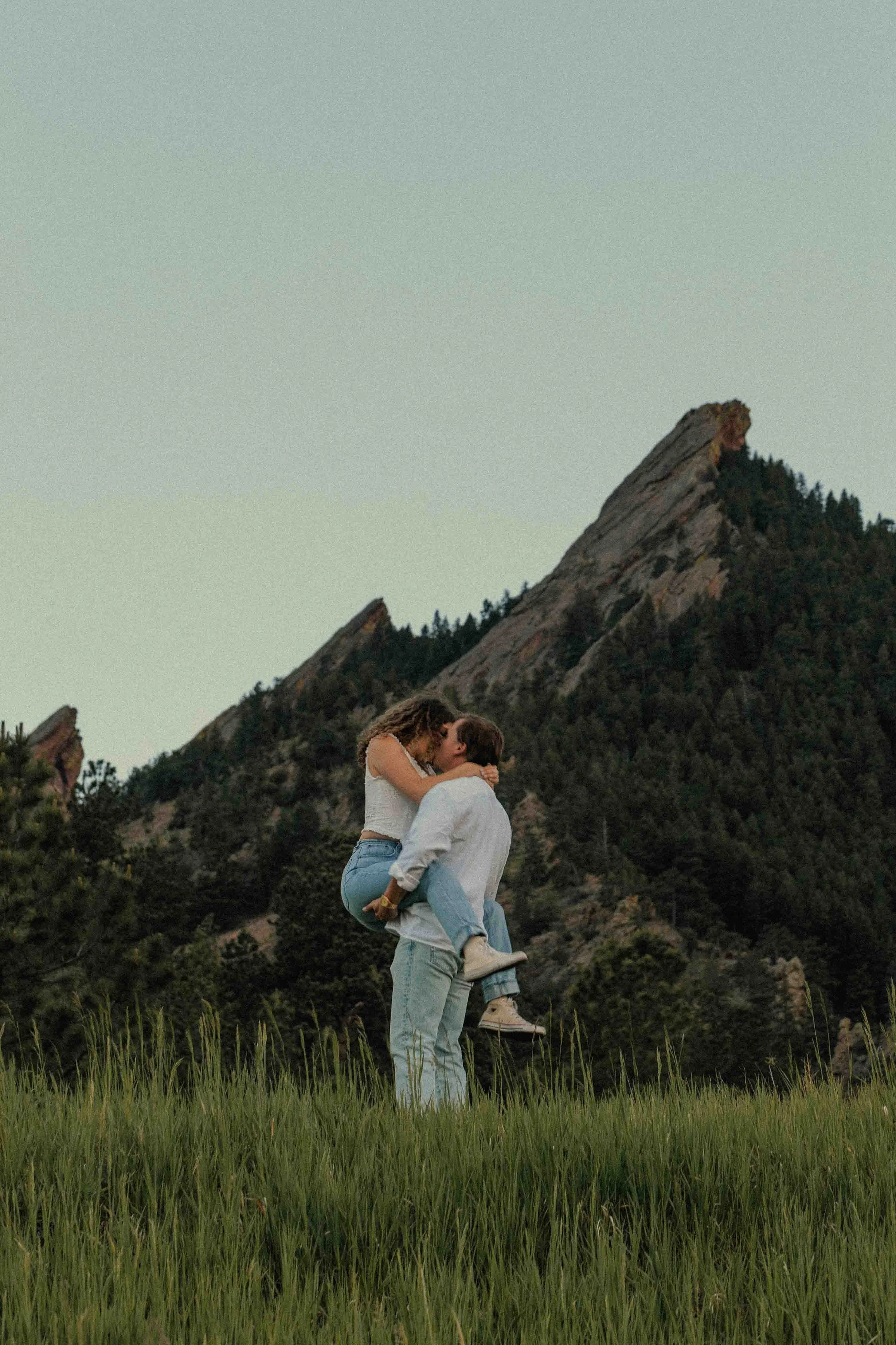 A couple embracing in a grassy field with mountains in the background at sunset