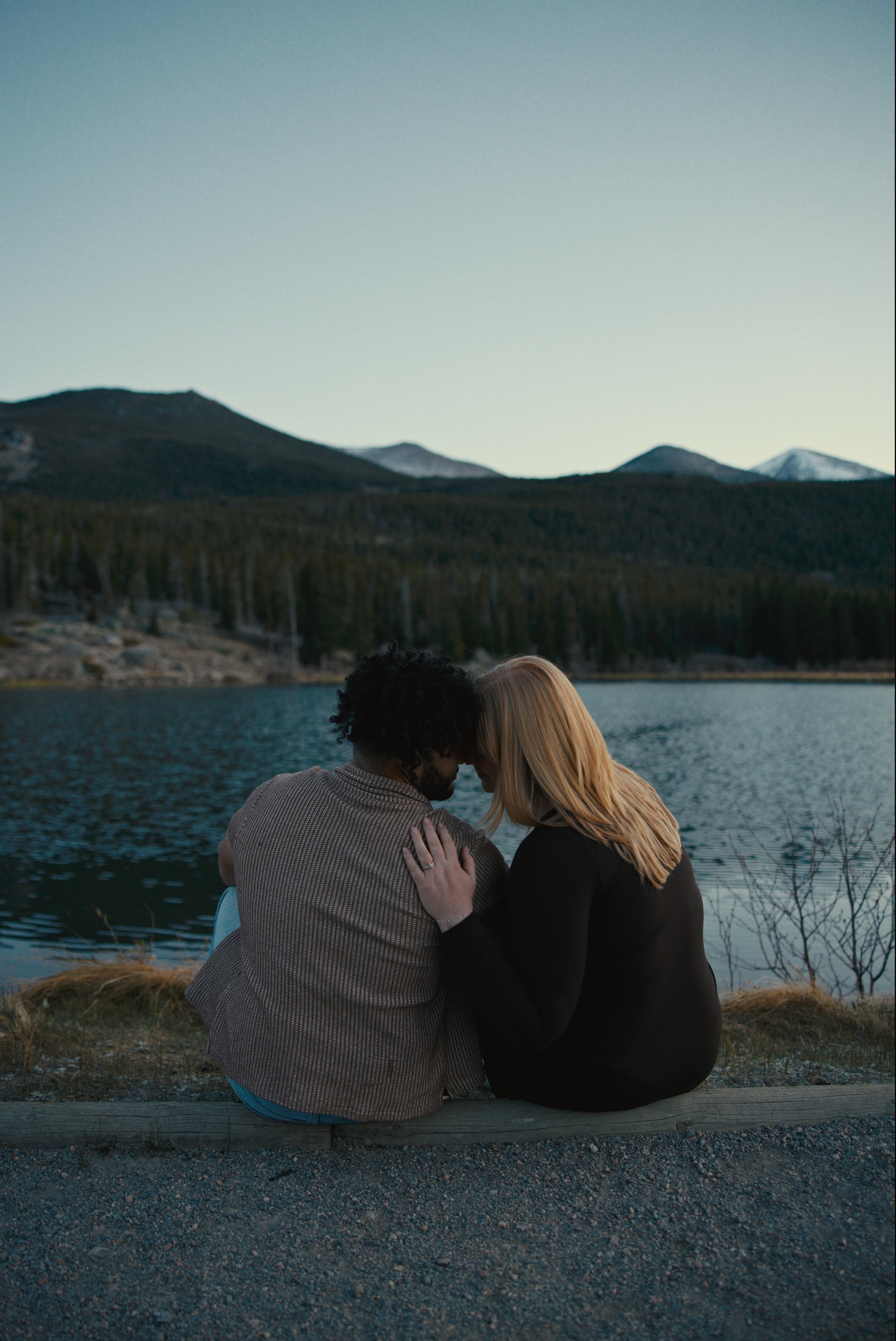 A couple sitting by a lake with mountains in the background, their foreheads touching in an intimate embrace during dusk.