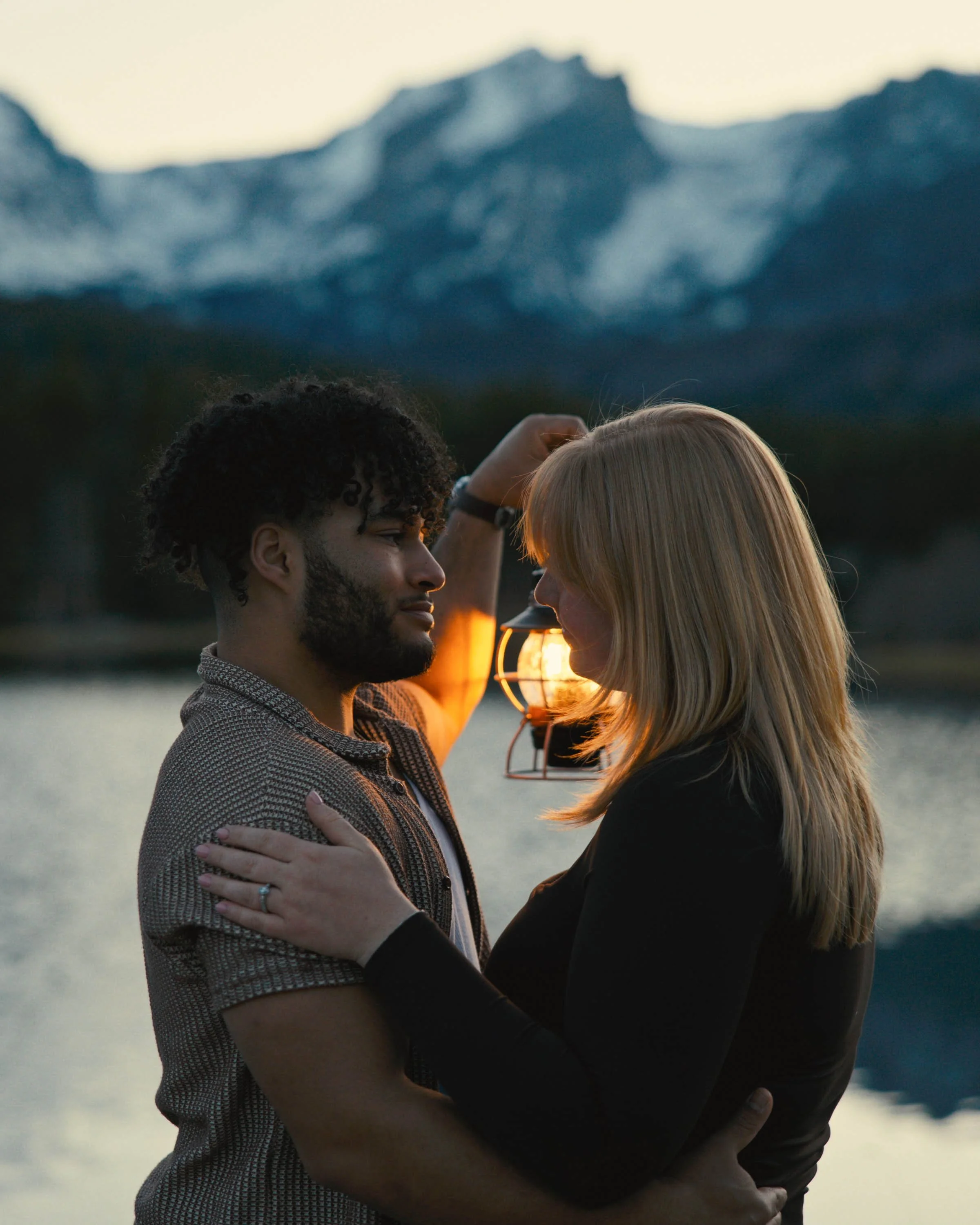 A couple faces each other closely at sunset by a body of water, with mountains in the background, holding each other affectionately.