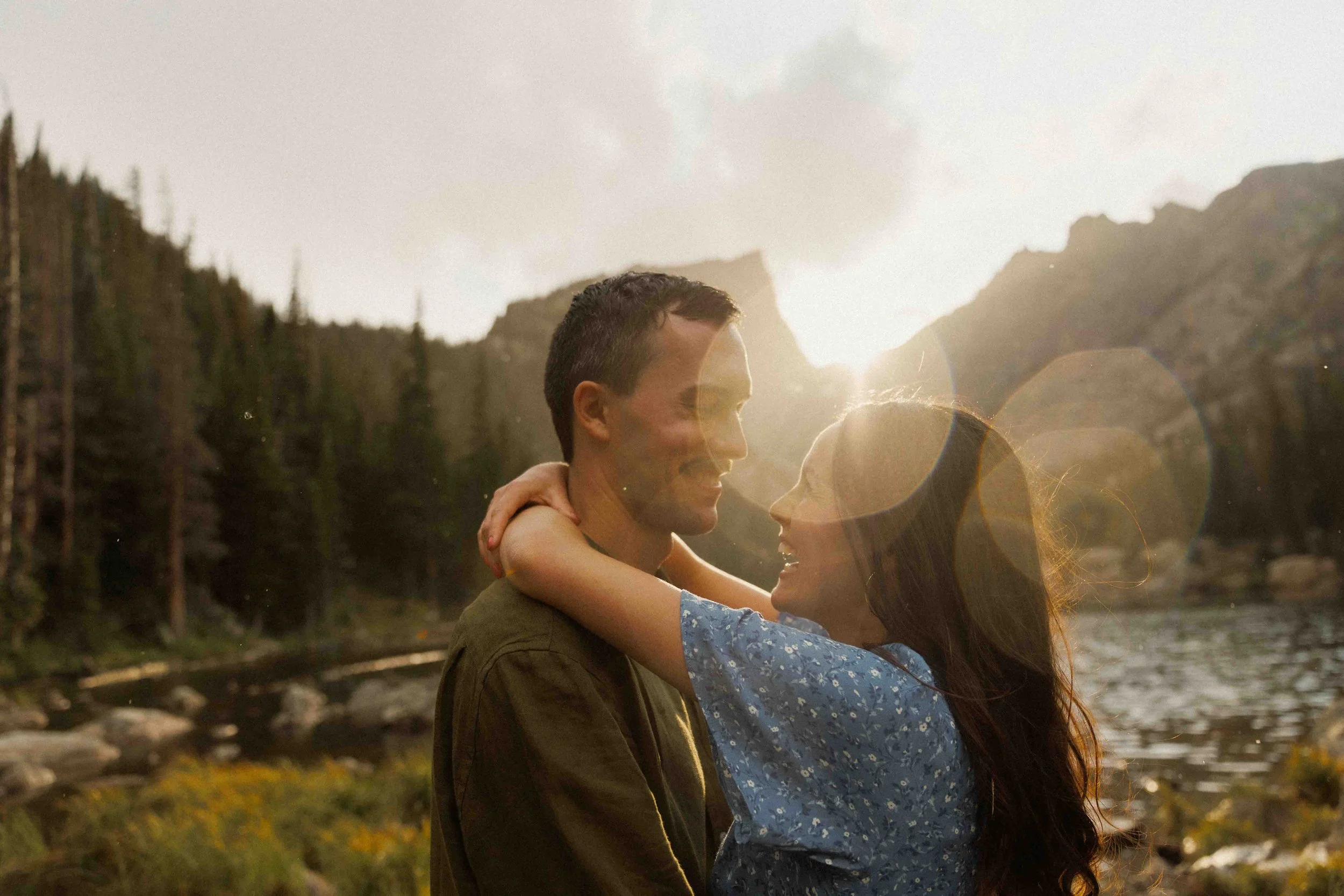 A couple embracing outdoors during sunset with mountain scenery in the background and lens flare effect.