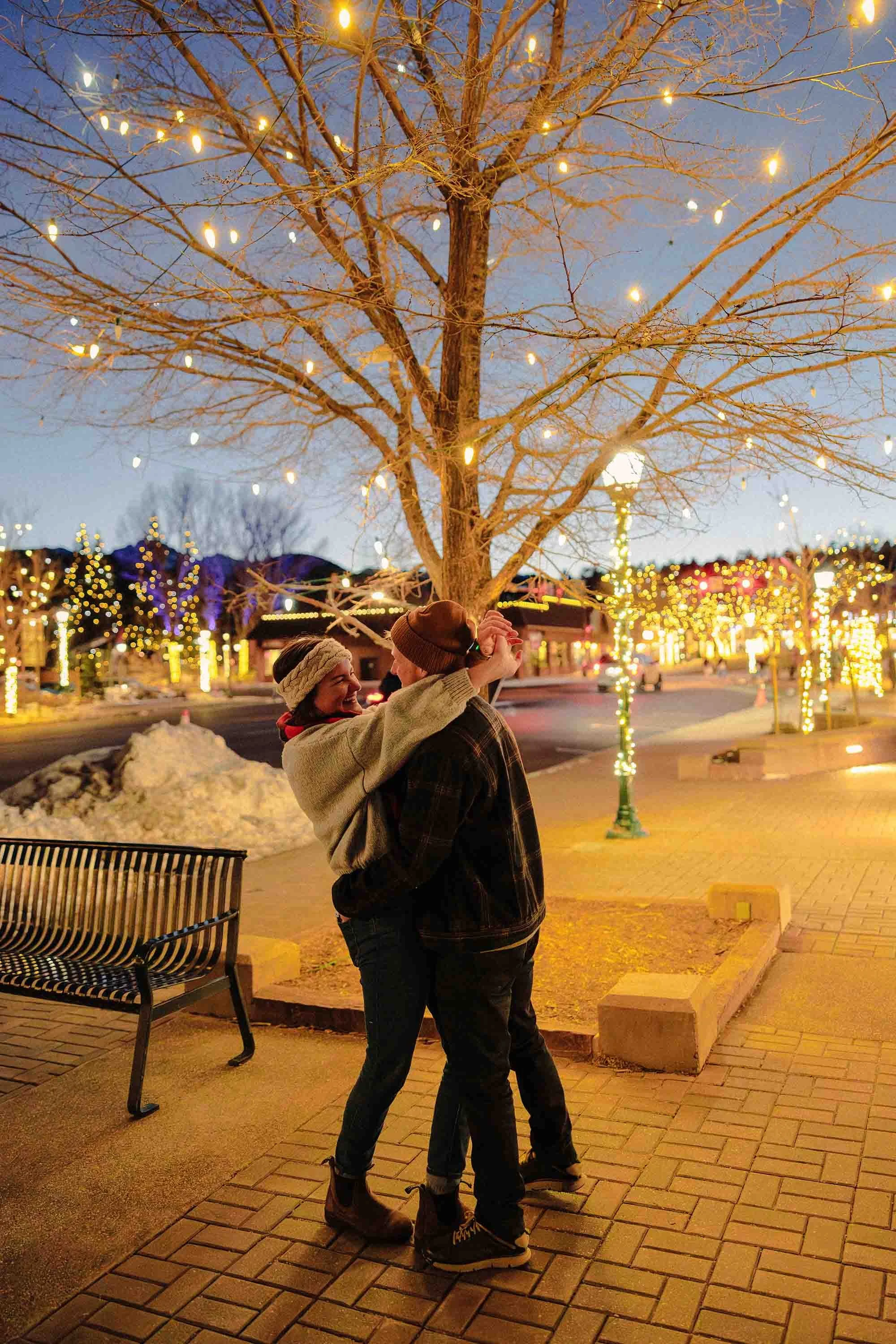 A couple is dancing and smiling under a lit, leafless tree decorated with string lights in a city park at night, with snow patches on the ground and warm yellow streetlights in the background.