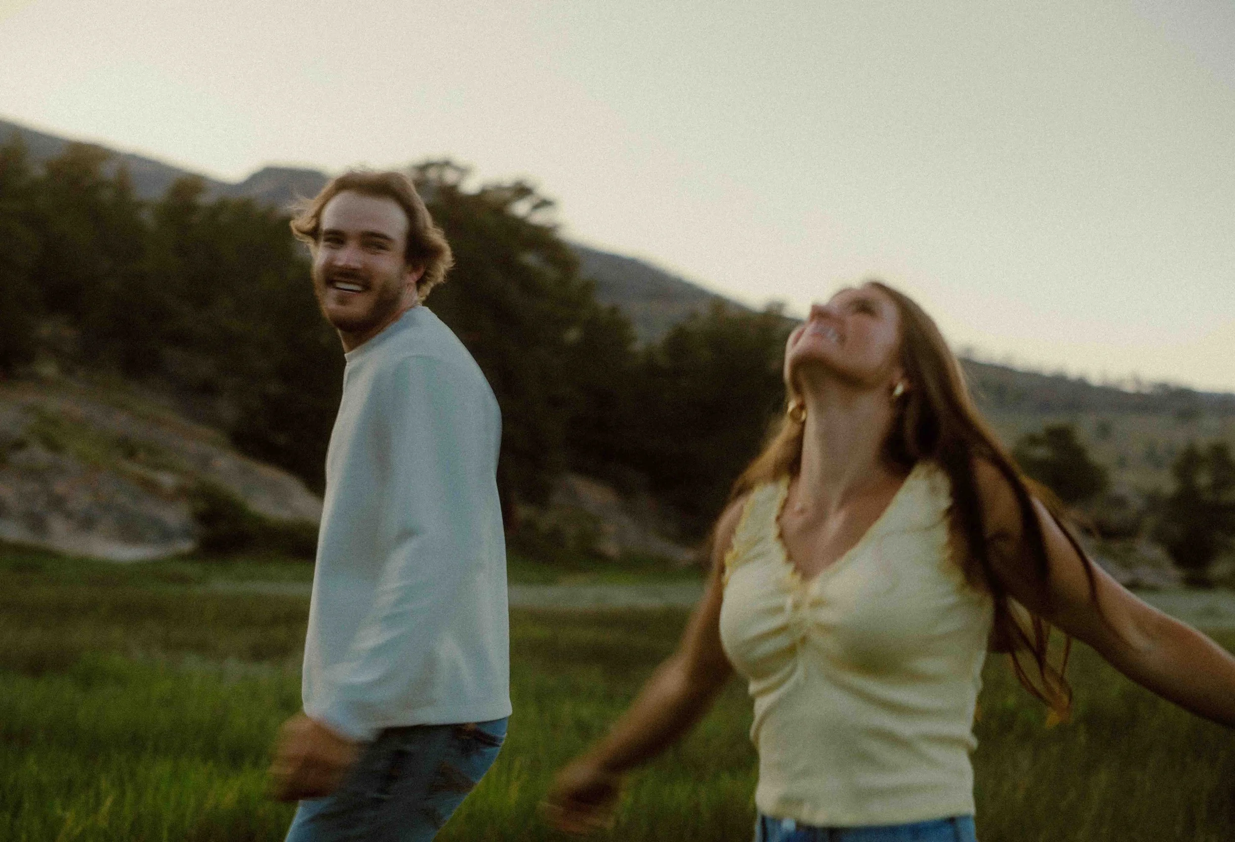 A young man and woman are outdoors, smiling and laughing as they walk through a grassy field with hills and trees in the background during sunset.