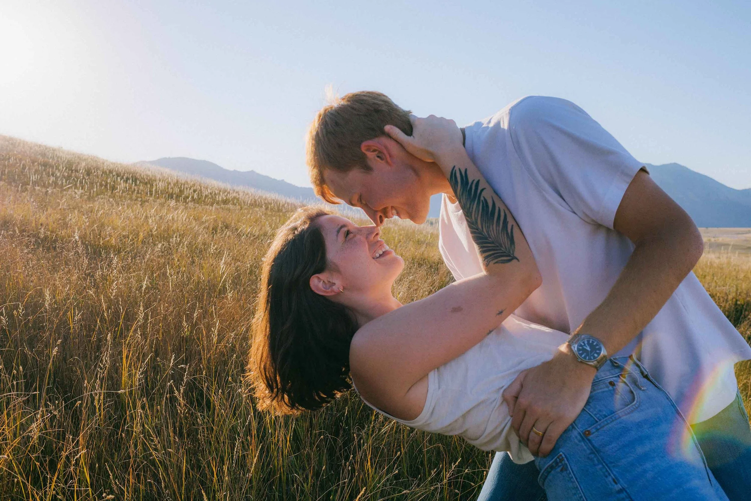 A couple sharing a romantic moment outdoors in a grassy field with mountains in the background, the man leaning over and touching foreheads with the woman, both smiling.